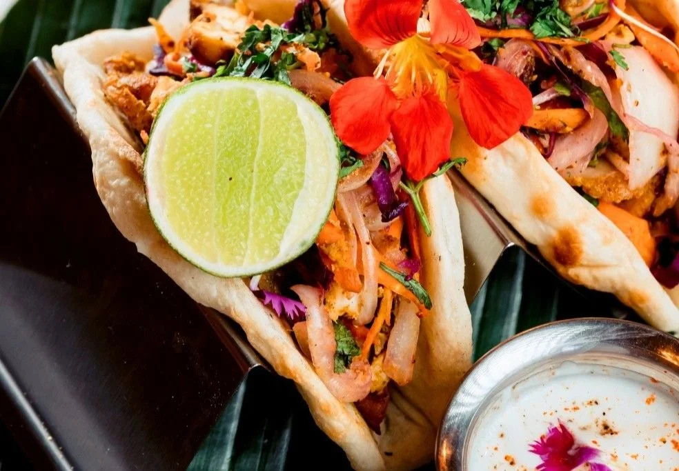 Two tandoori chicken tacos topped with fresh herbs, shredded vegetables, and a lime wedge, served on a black plate.