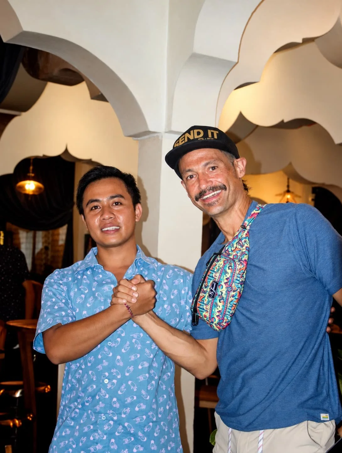 Two men shaking hands, one wearing a blue patterned shirt and the other in a blue t-shirt with a colorful bag and a black cap with 'SEND IT' written on it, in a cozy indoor setting.