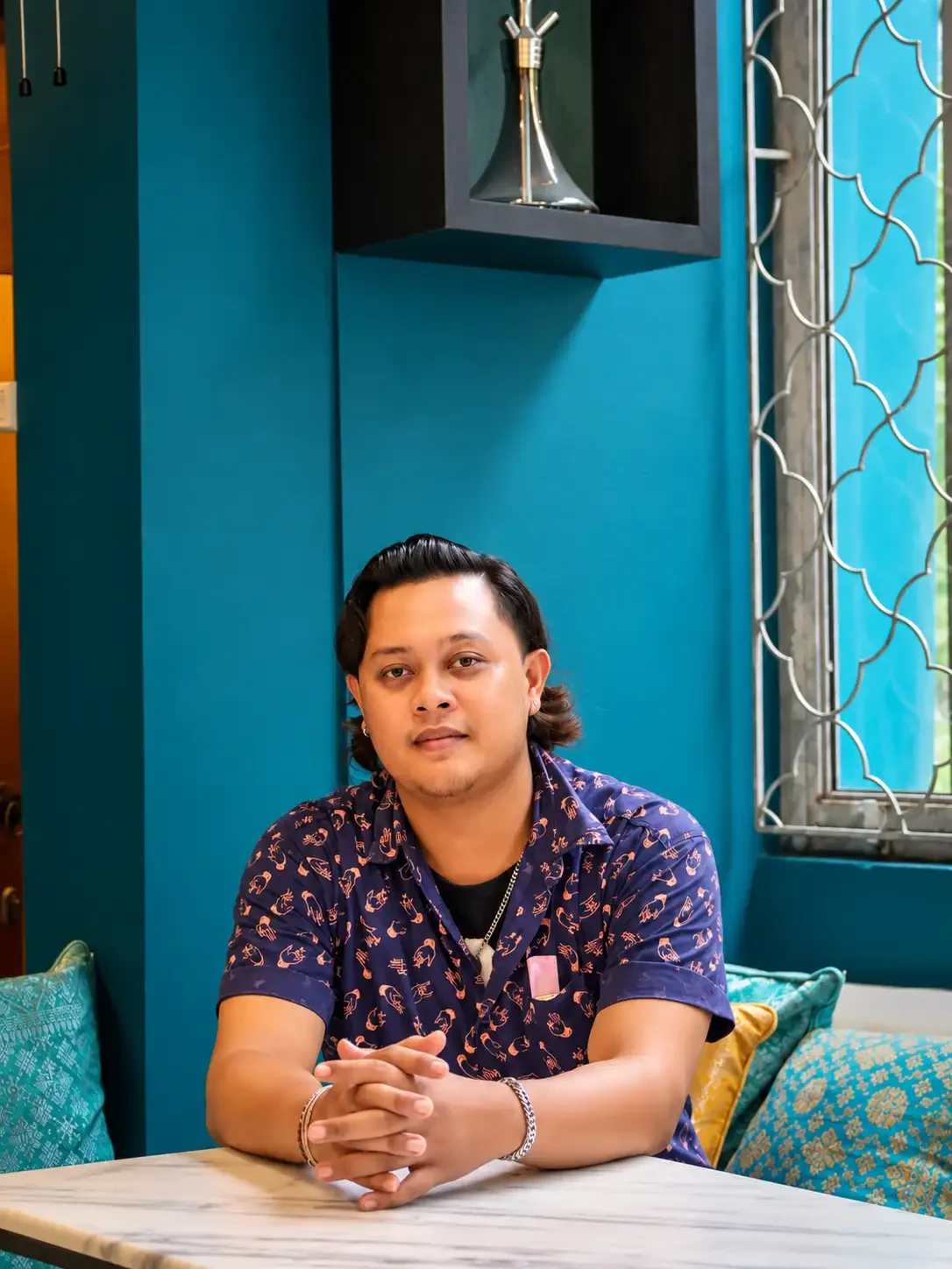 A young man with black hair sitting at a table with his hands clasped, wearing a navy blue shirt with a pattern, in a room with teal walls and decorative pillows, next to a window with geometric metal grillwork.
