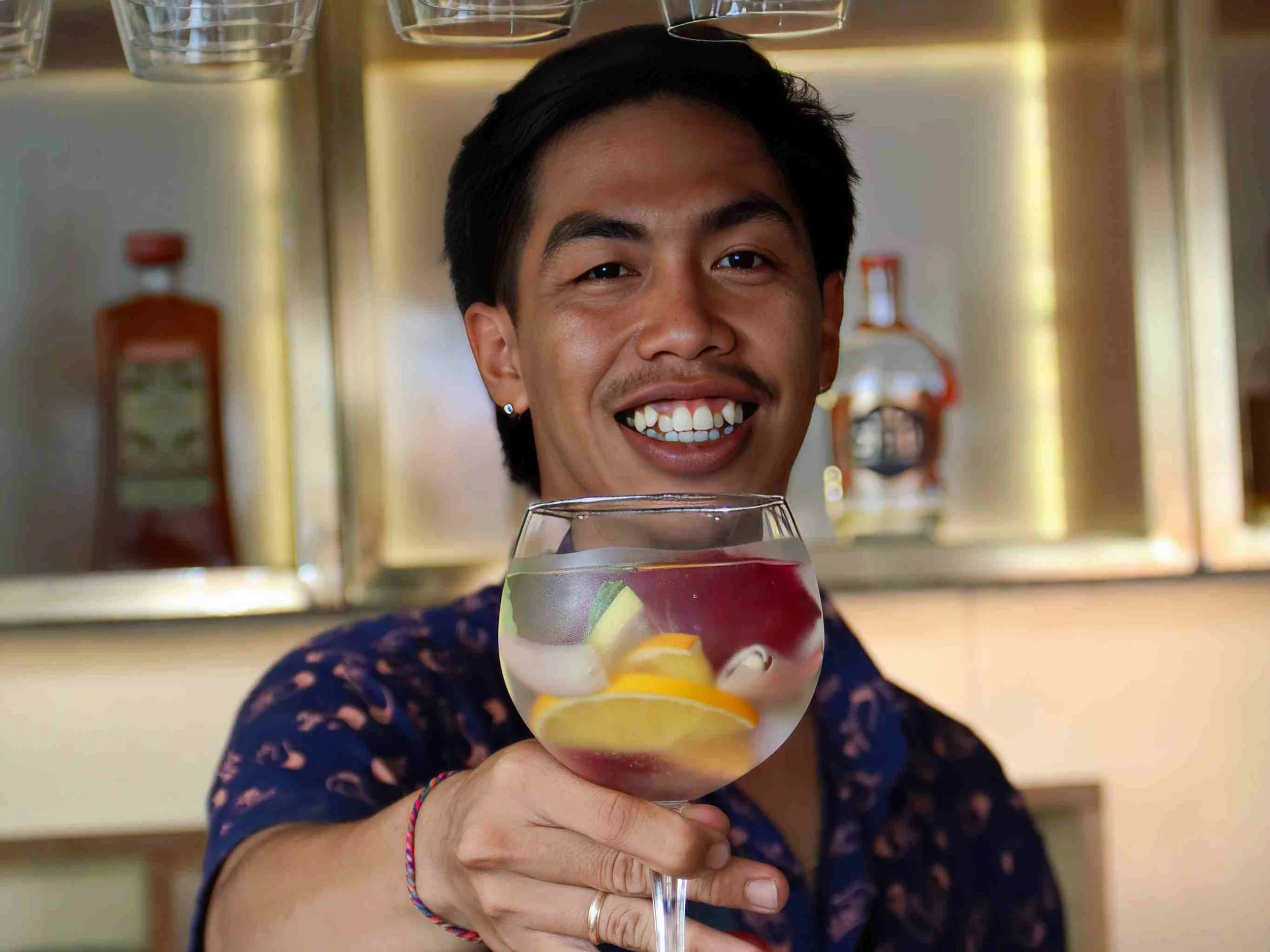 A person smiling and holding a glass of cocktail with lemon and lime slices inside.