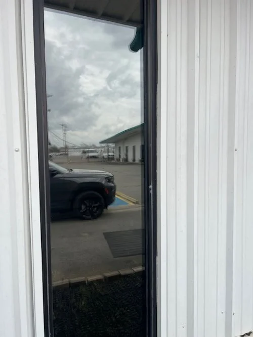 Reflection of a cloudy sky and parked car in a glass door, with a white metal building exterior.