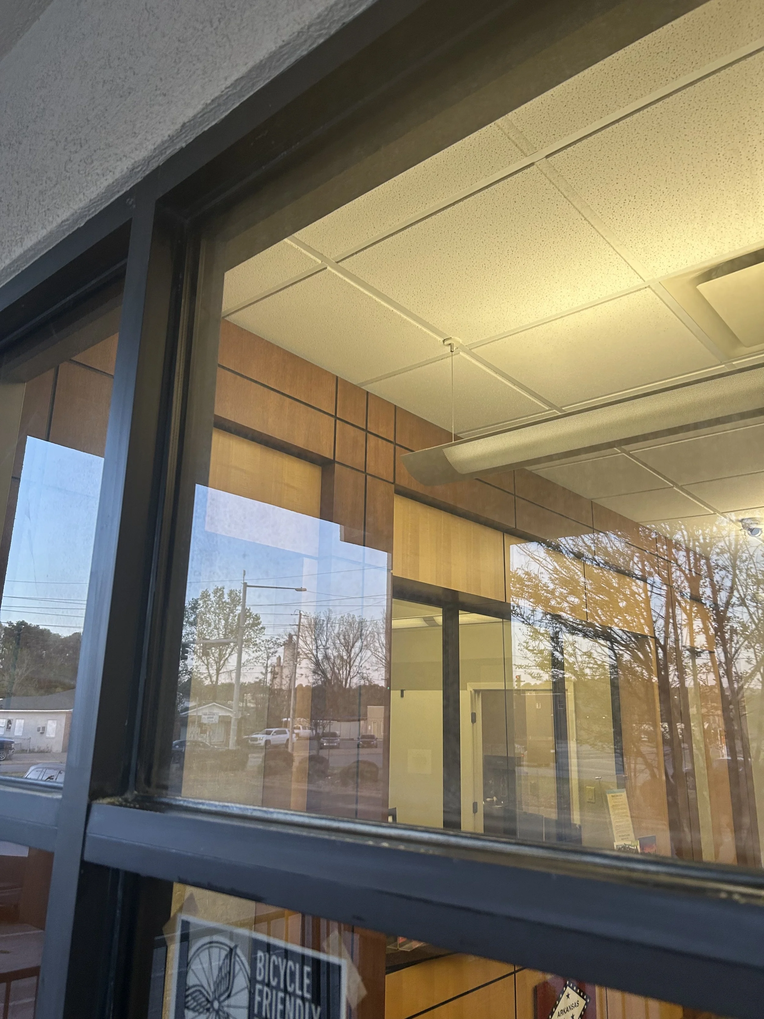 View through a large glass window showing the interior of a building with wood paneling, drop ceiling, fluorescent light, and reflections of trees and cars outside.