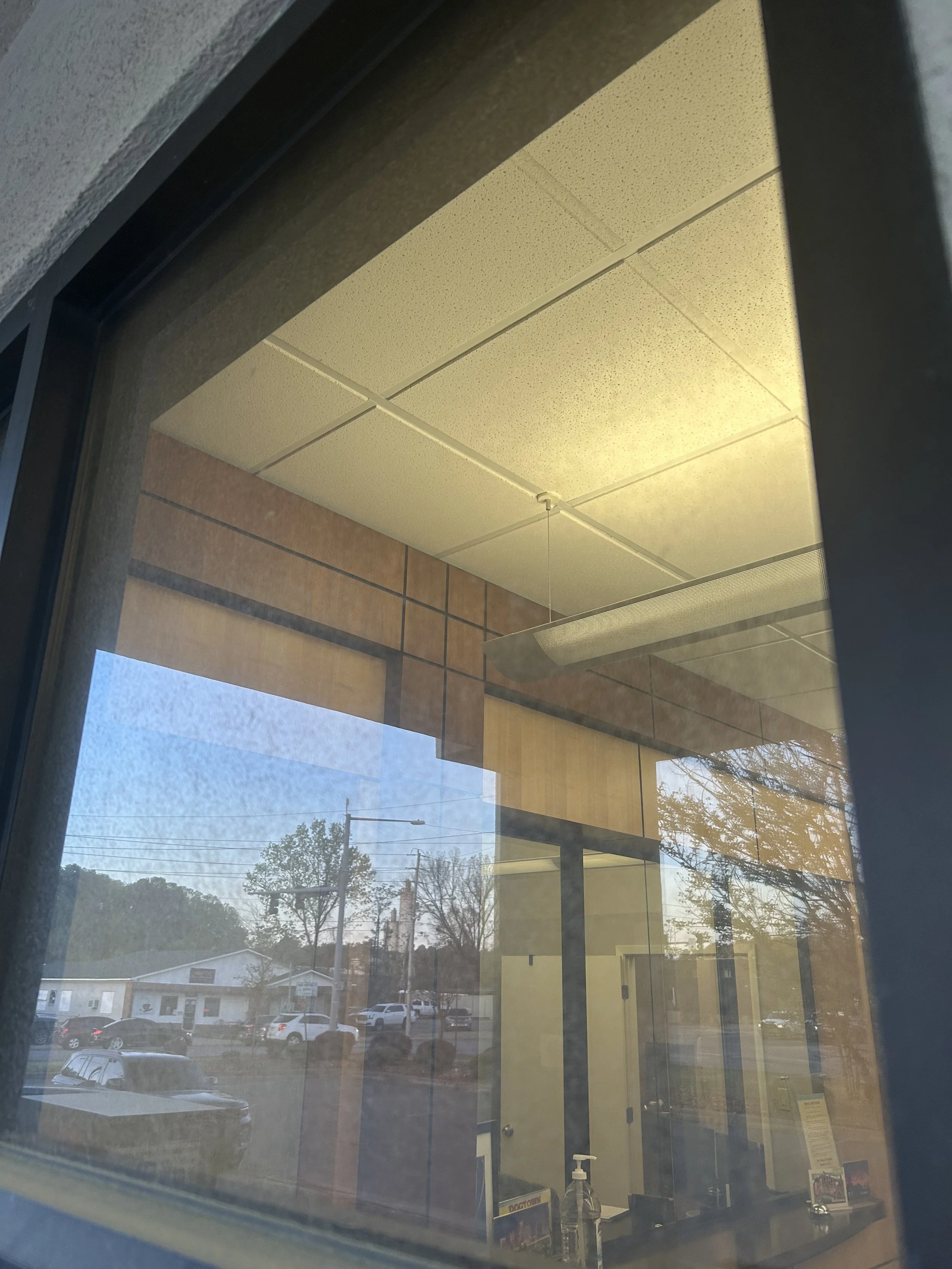 Photo taken from outside a window showing the interior ceiling with a fluorescent light, a door, and a view of parked cars and trees outside.