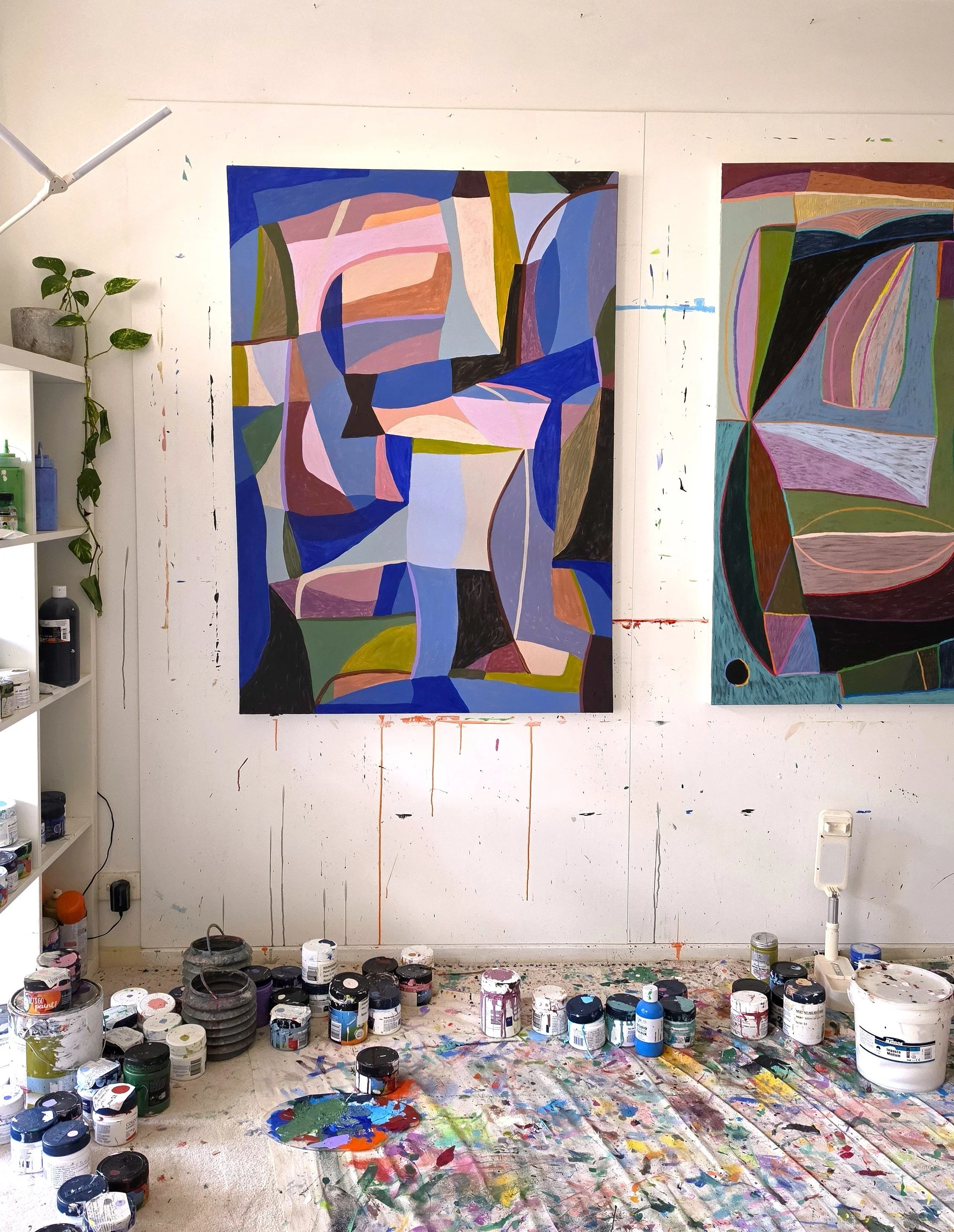 Art studio with two large abstract paintings hanging on the wall, paint supplies including numerous jars of paint on the floor and shelves, and a paint palette with mixed colors on the floor, all in a workspace with splattered paint on the floor and wall.