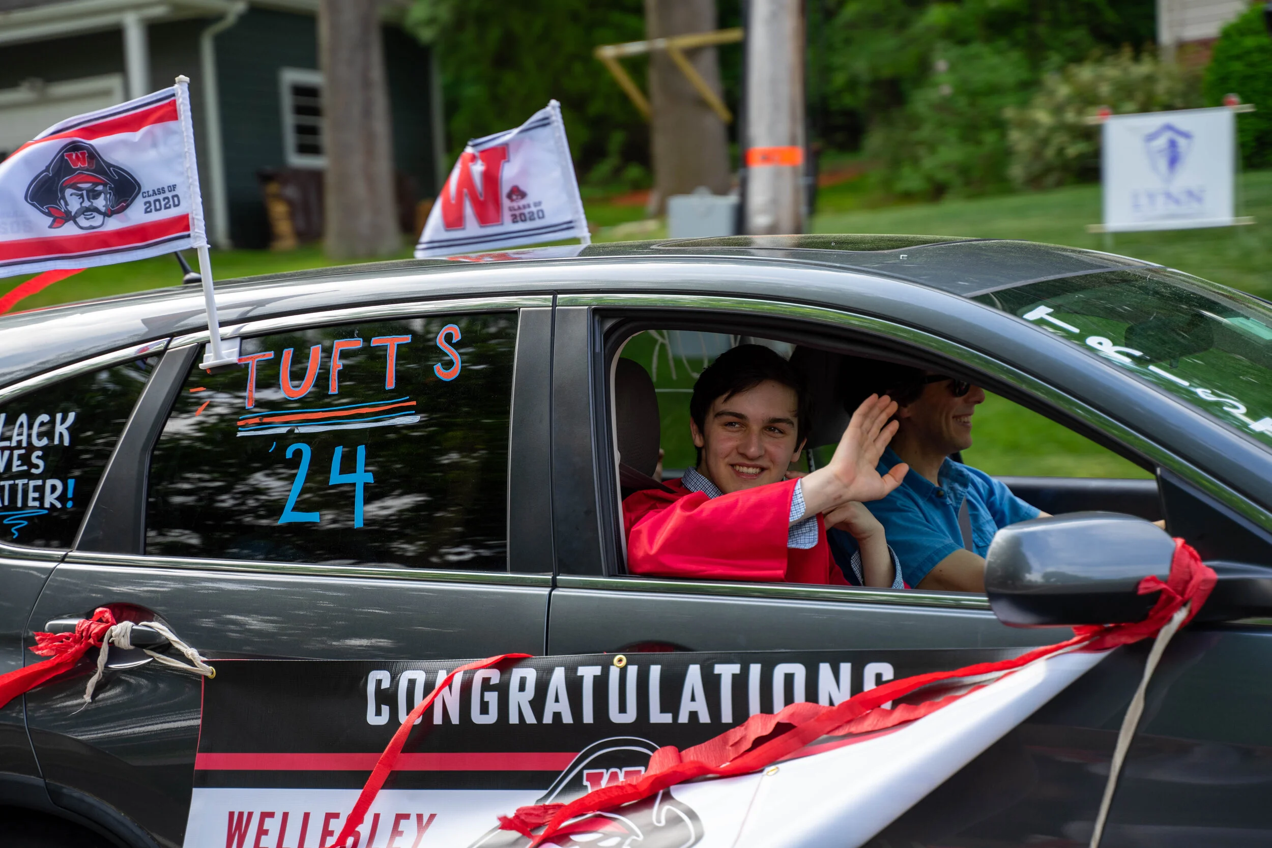 A decorated car with flags and banners celebrating graduation, with a young man and a woman inside, smiling and waving, in a neighborhood setting with trees and a house in the background.