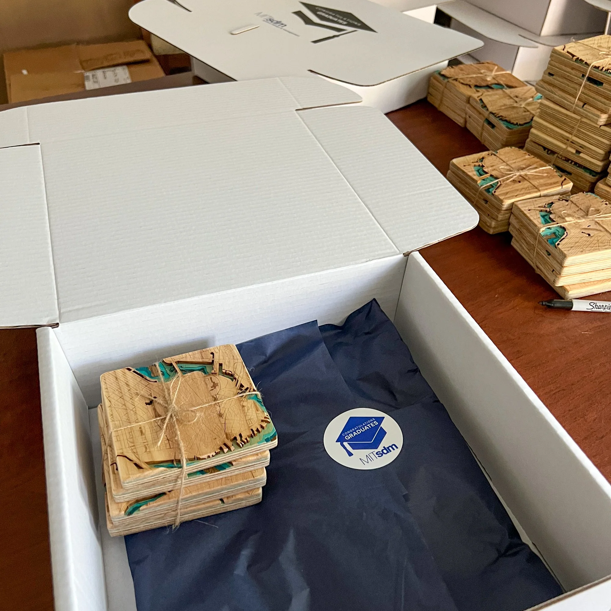 A cardboard box with a white exterior and an open top on a dark blue surface, next to a stack of smaller, similarly decorated boxes with blue and orange designs tied with string. In the background, there are white shelves and an assortment of brown b