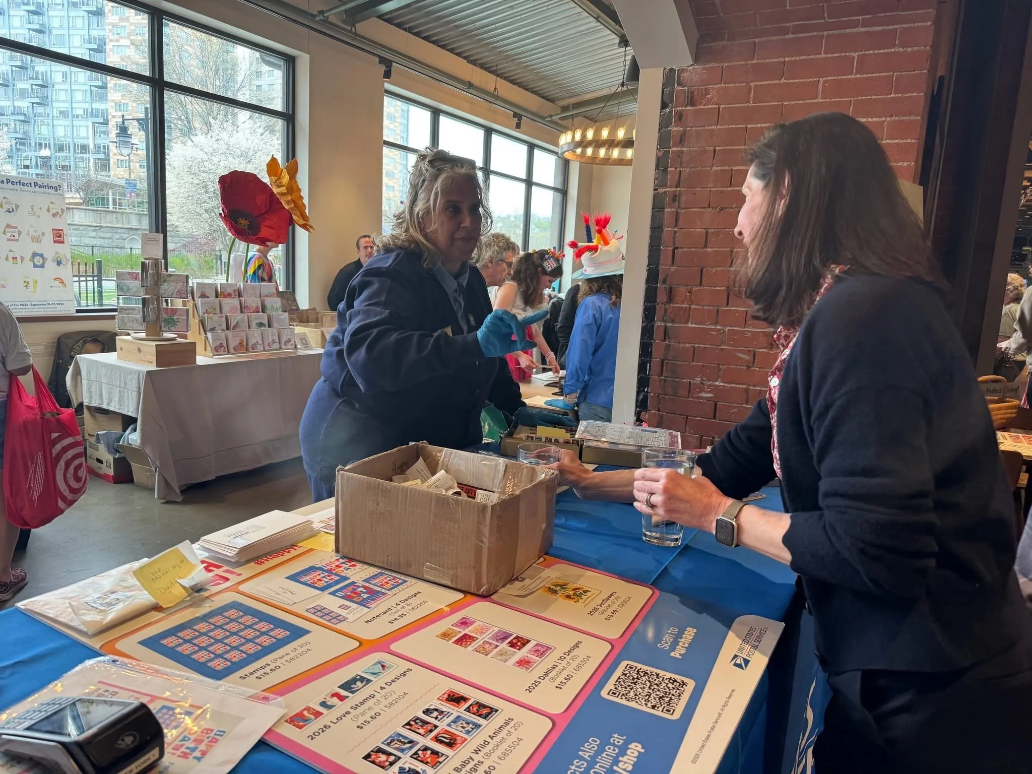 People at a table with lottery tickets, promotional materials, and a cash box inside a modern building with large windows and a brick wall.