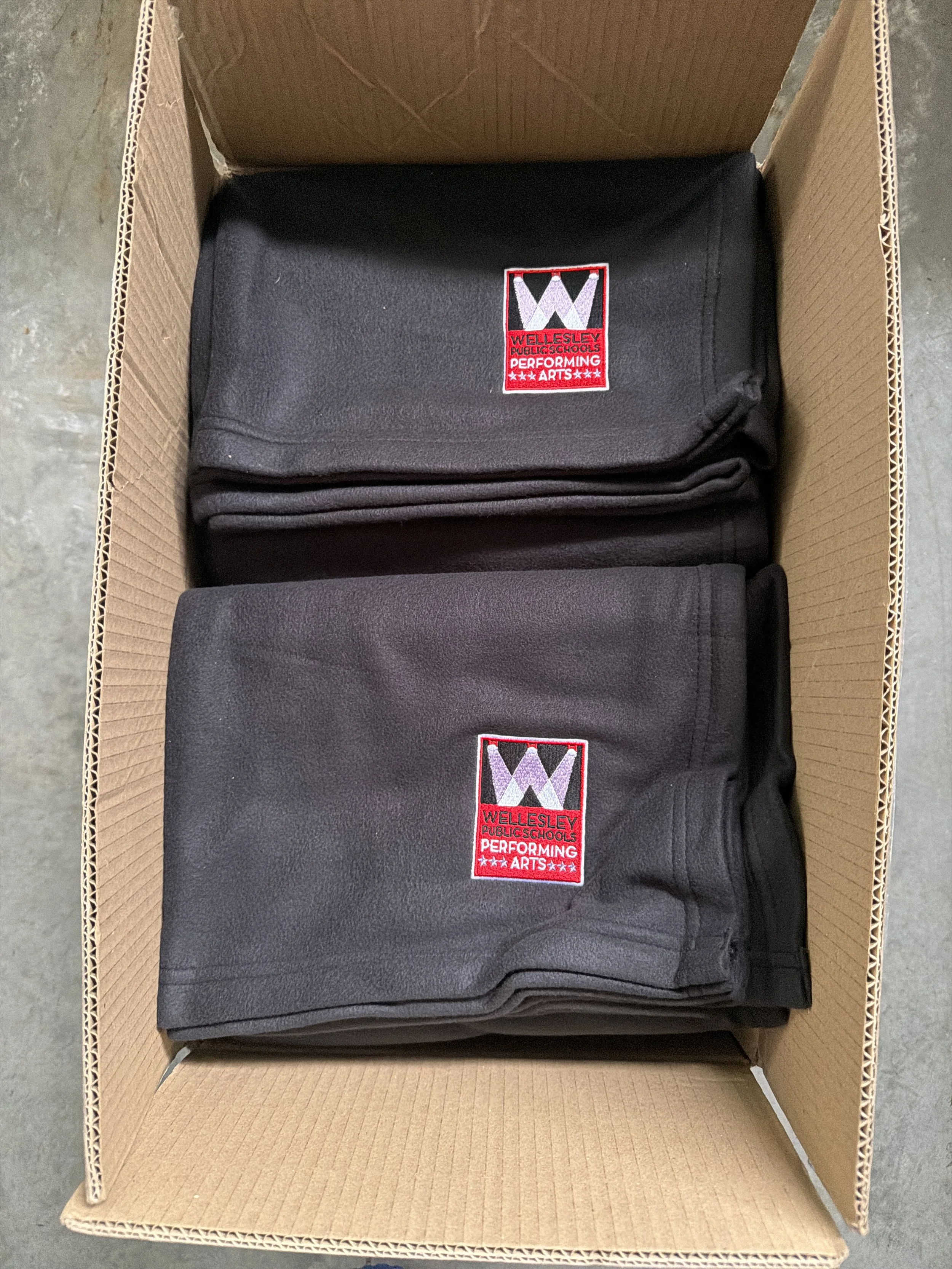 A cardboard box containing multiple folded black uniforms or pants with patches that read "Wellesley Public Schools Performing Arts".