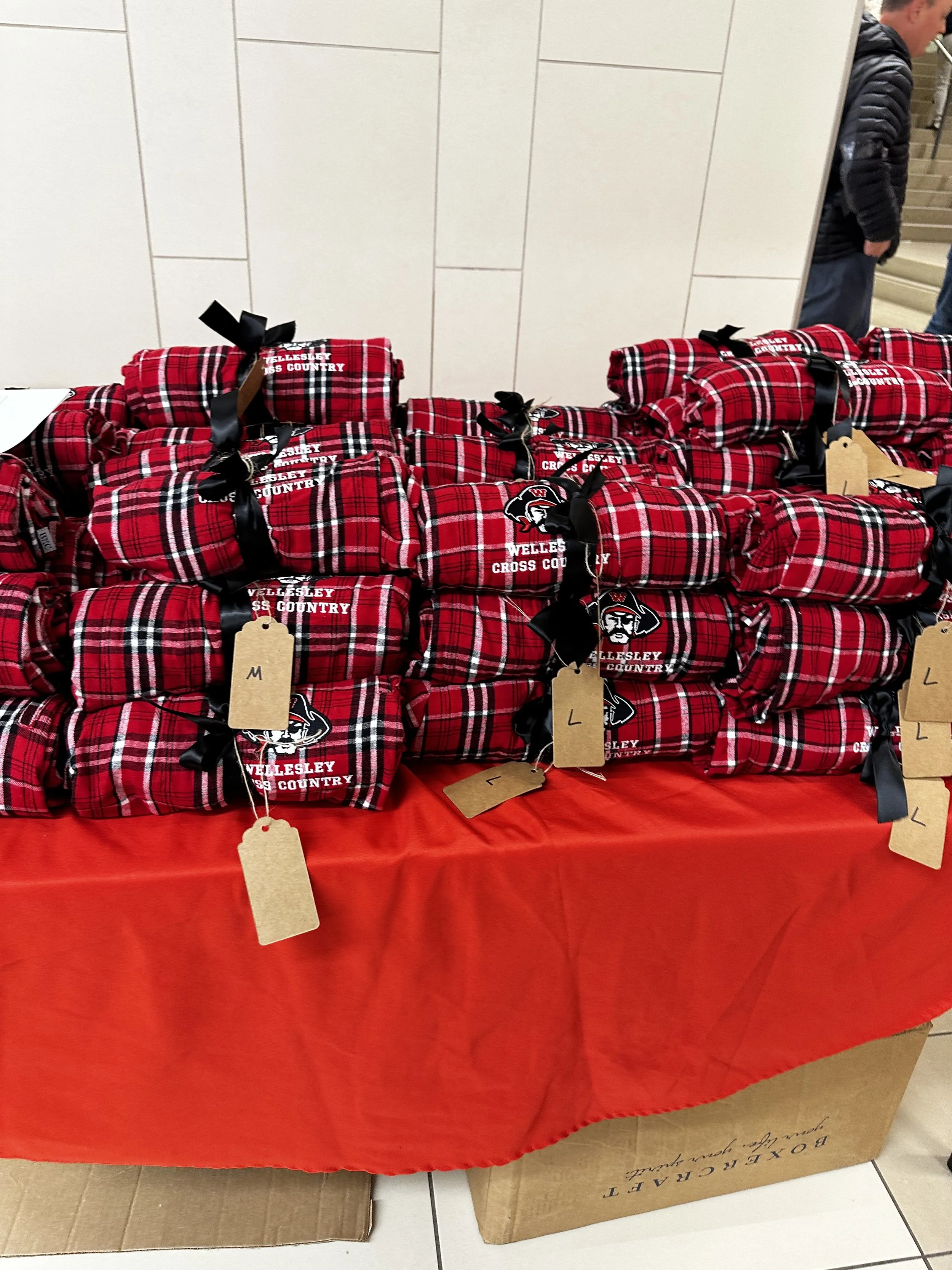 Stacked red plaid blankets with black ribbons and tags on a red tablecloth, some with a logo and text, beside large cardboard boxes at a retail or event setting.