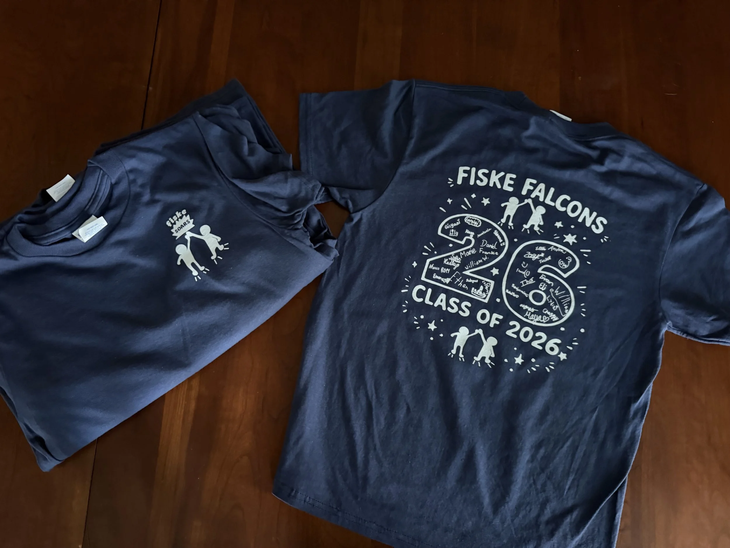 Two dark blue T-shirts with white text and graphics laid out on a wooden surface. The shirts are associated with a class of 2026 celebration, featuring illustrations of children holding hands and names of students.