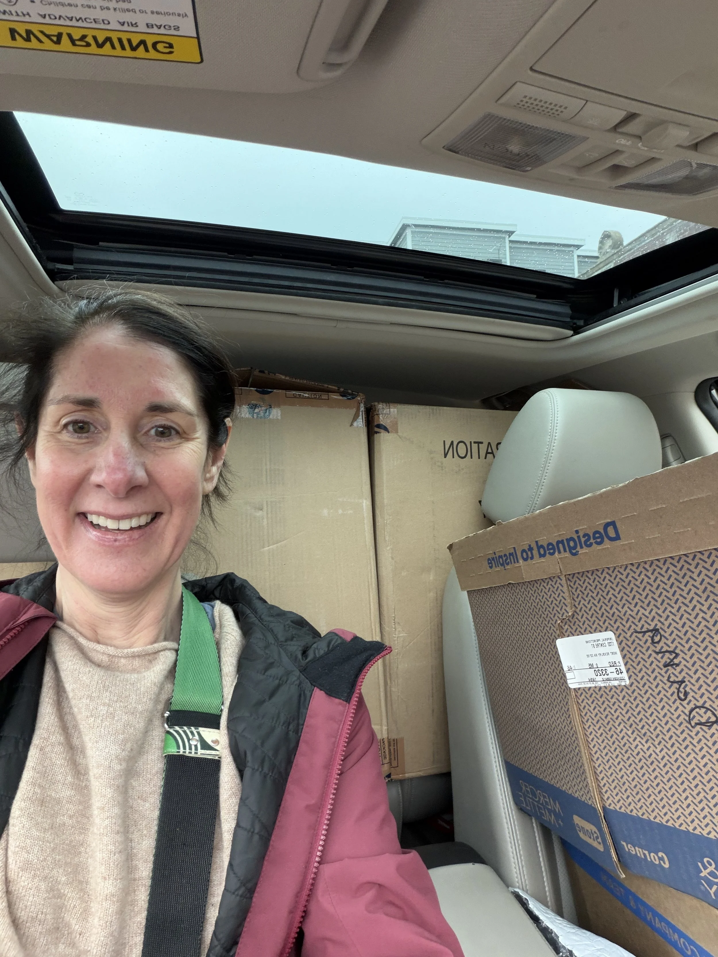 A woman smiling inside a car with a sunroof, packed with boxes and packages, including beer and wine boxes.