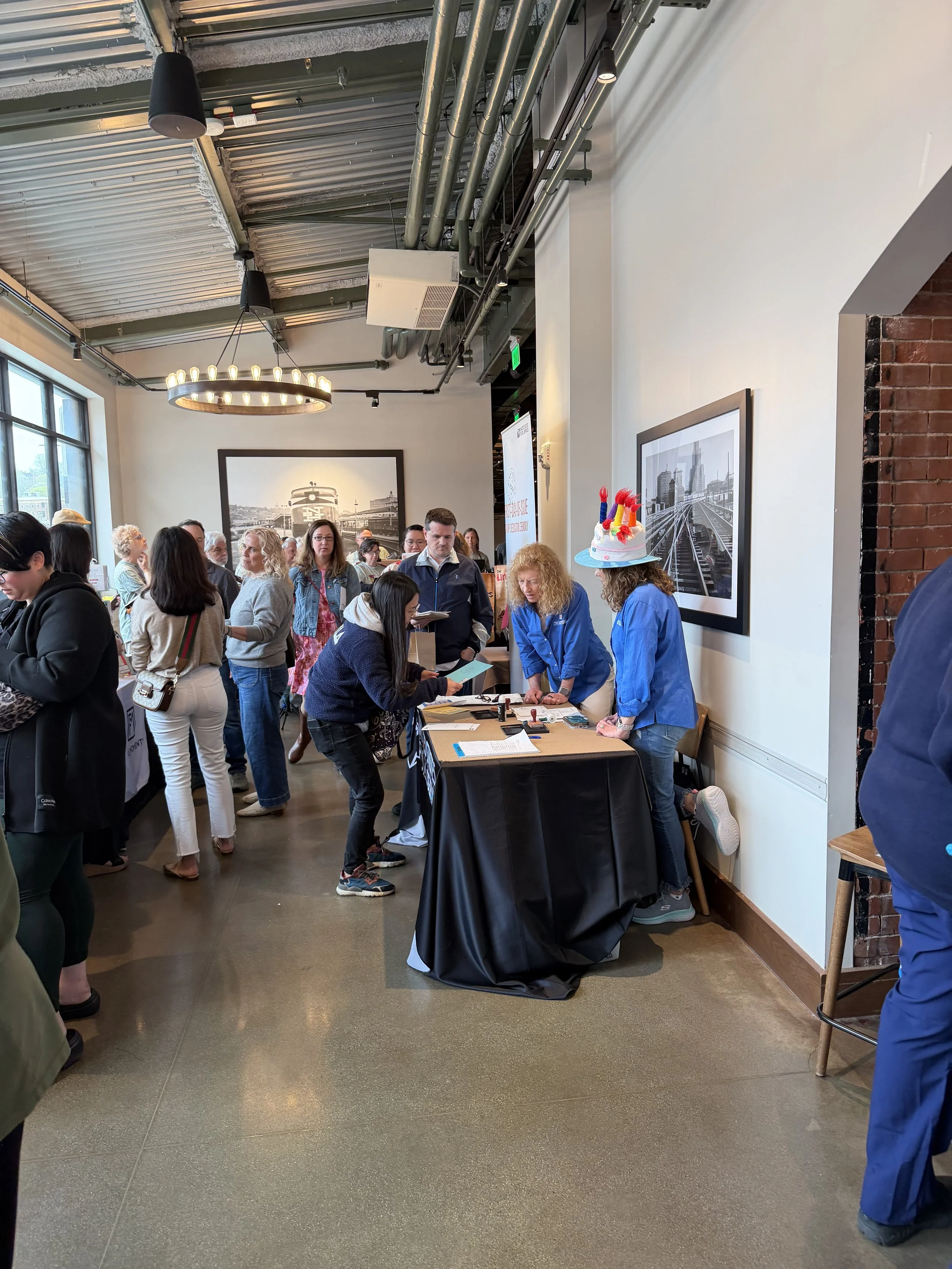 People gathered in a modern indoor space, some standing around a table with a black tablecloth, while two women wear a patriotic birthday hat with red, white, and blue decorations. The room features large windows, framed black and white photos of tra