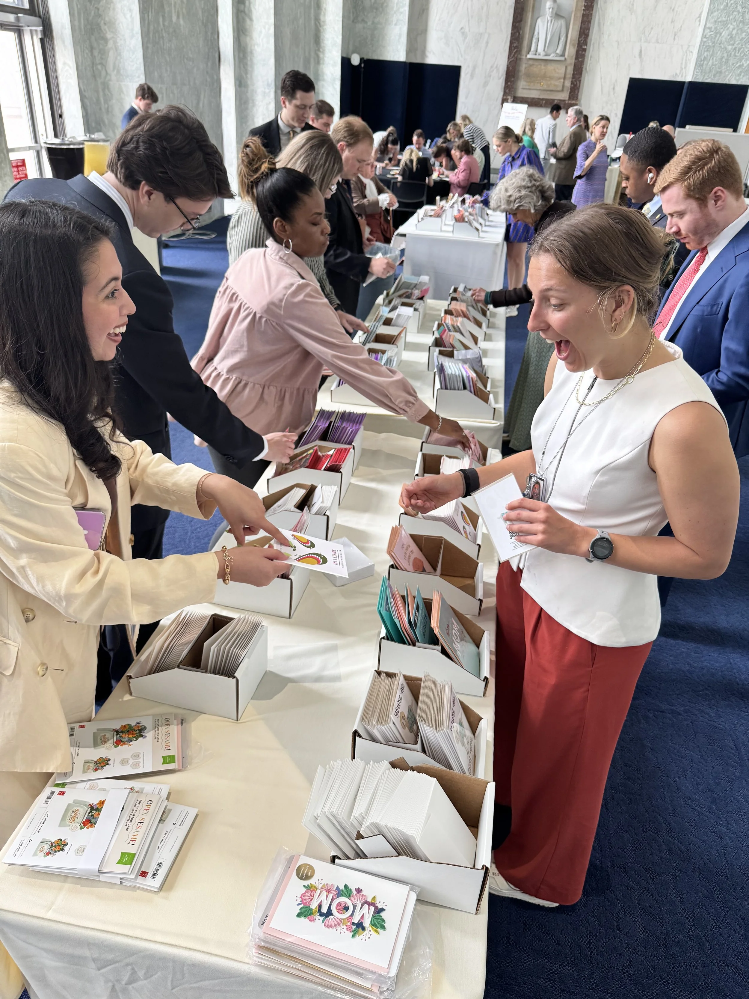 People browsing and selecting greeting cards at a display table in a large indoor event space.