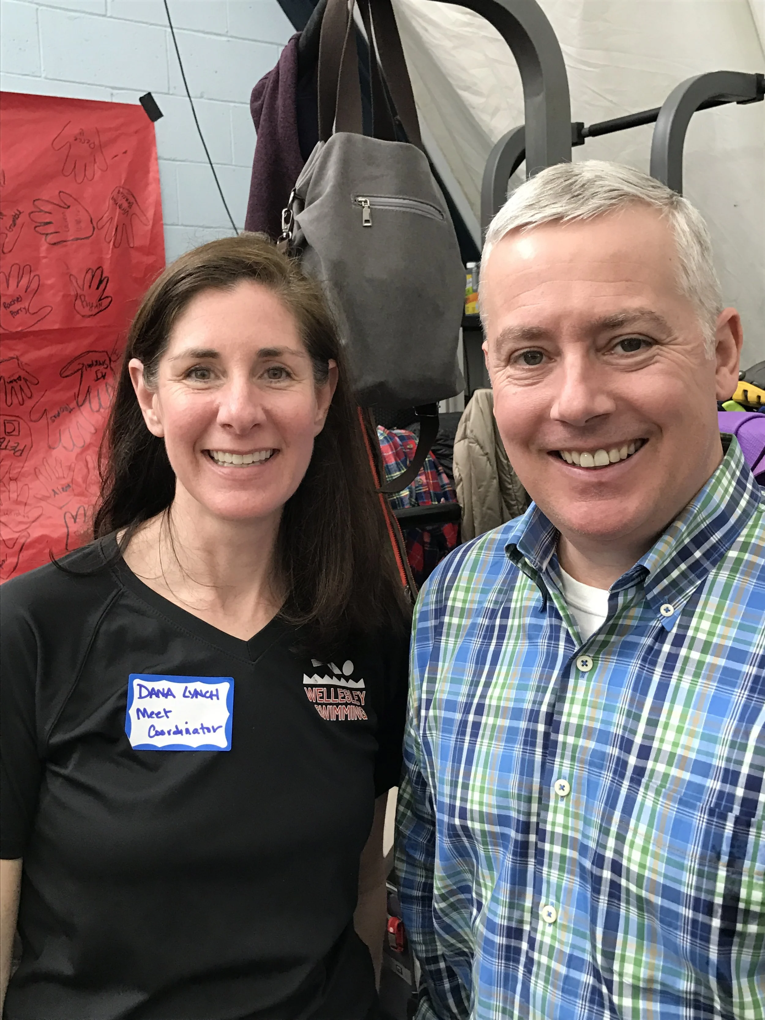 A woman with dark brown hair wearing a black jacket and a blue name tag and a man with gray hair wearing a colorful plaid shirt smiling for a selfie. Behind them are backpacks, clothing, and a white brick wall with a red poster with handprints.
