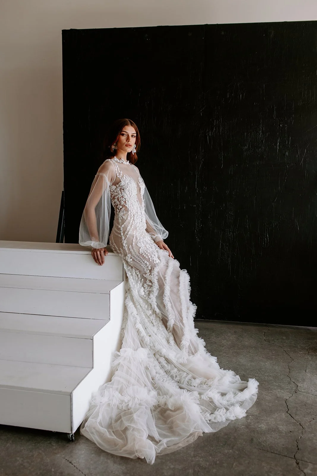 Woman in a white lace wedding gown with puffed sleeves sitting on white staircase, against a black background.
