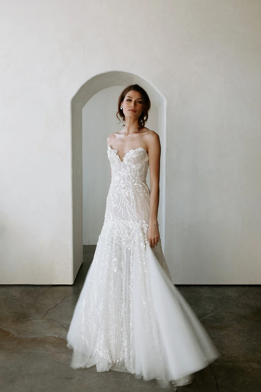 A woman in a white, strapless wedding gown with intricate beading stands in front of an arched doorway against a light-colored wall.