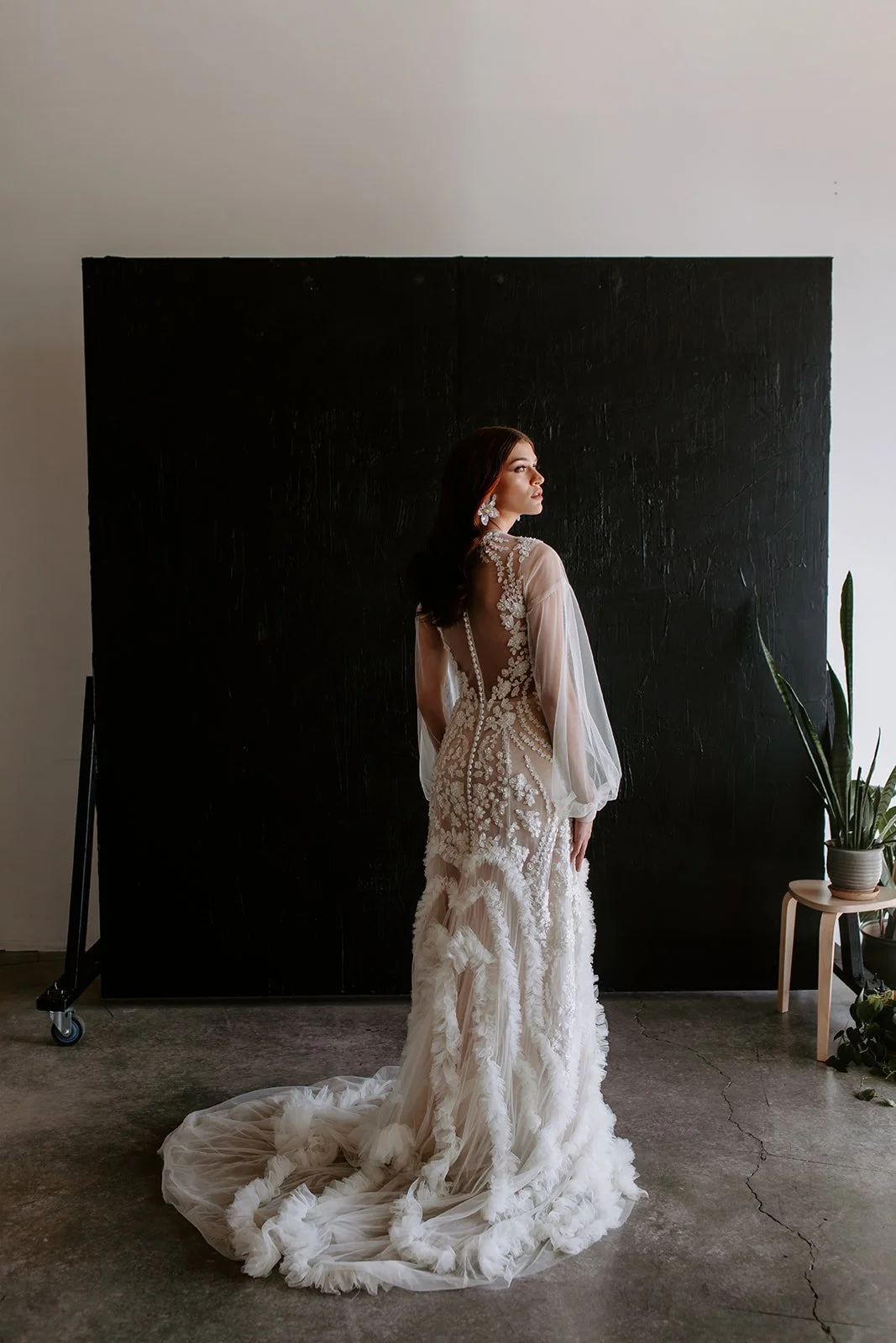 Woman in an elegant white wedding dress with floral lace details, posing in front of a black background, with a potted plant on a stool nearby.