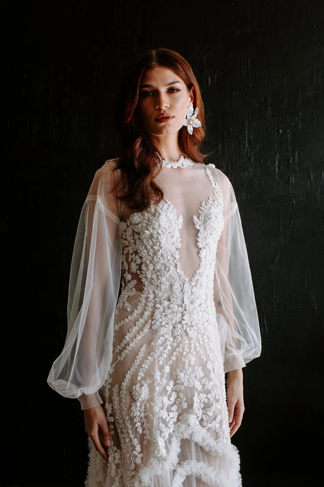Woman in an ornate white dress with floral embroidery and sheer sleeves, wearing large floral earrings, against a dark background.