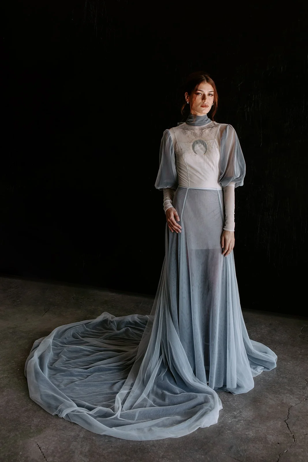 Woman wearing a flowing, light blue formal gown with sheer fabric and puffed sleeves, standing against a dark background.