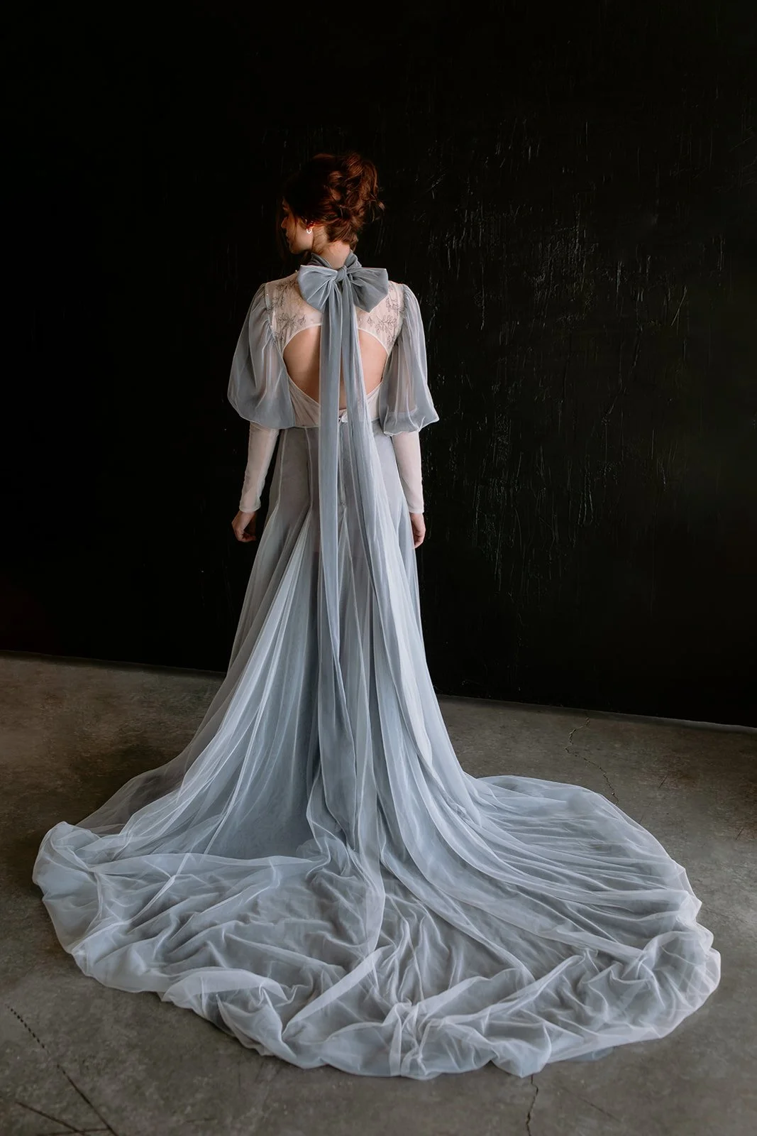 Person wearing a long, elegant, light gray dress with a bow on the back and sheer cape-like sleeves, standing against a dark background.