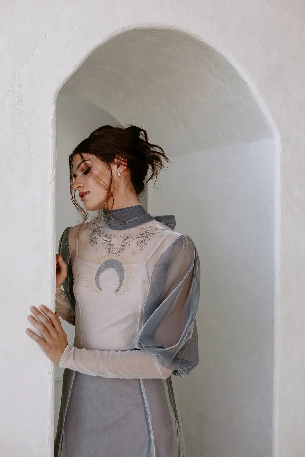 A woman in an elegant, sheer gown with a moon design, standing in an arched alcove with white walls.