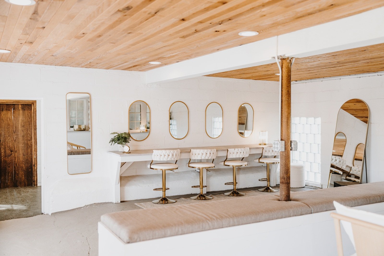 Interior of a modern, minimalist space with a white brick wall, a wooden ceiling, and a row of five white barstools with gold legs in front of a white countertop and five mirrors with gold frames. There is a wooden door on the left and a full-length 