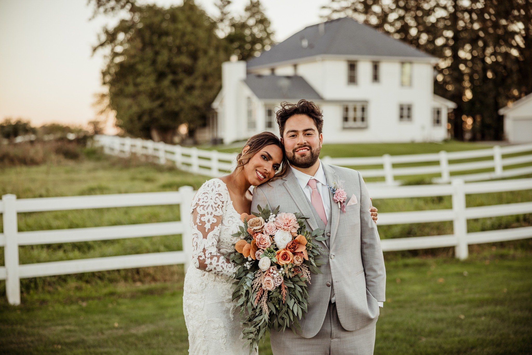 Bride and groom smiling outdoors with a white fence and house in the background, bride holding a bouquet of flowers.