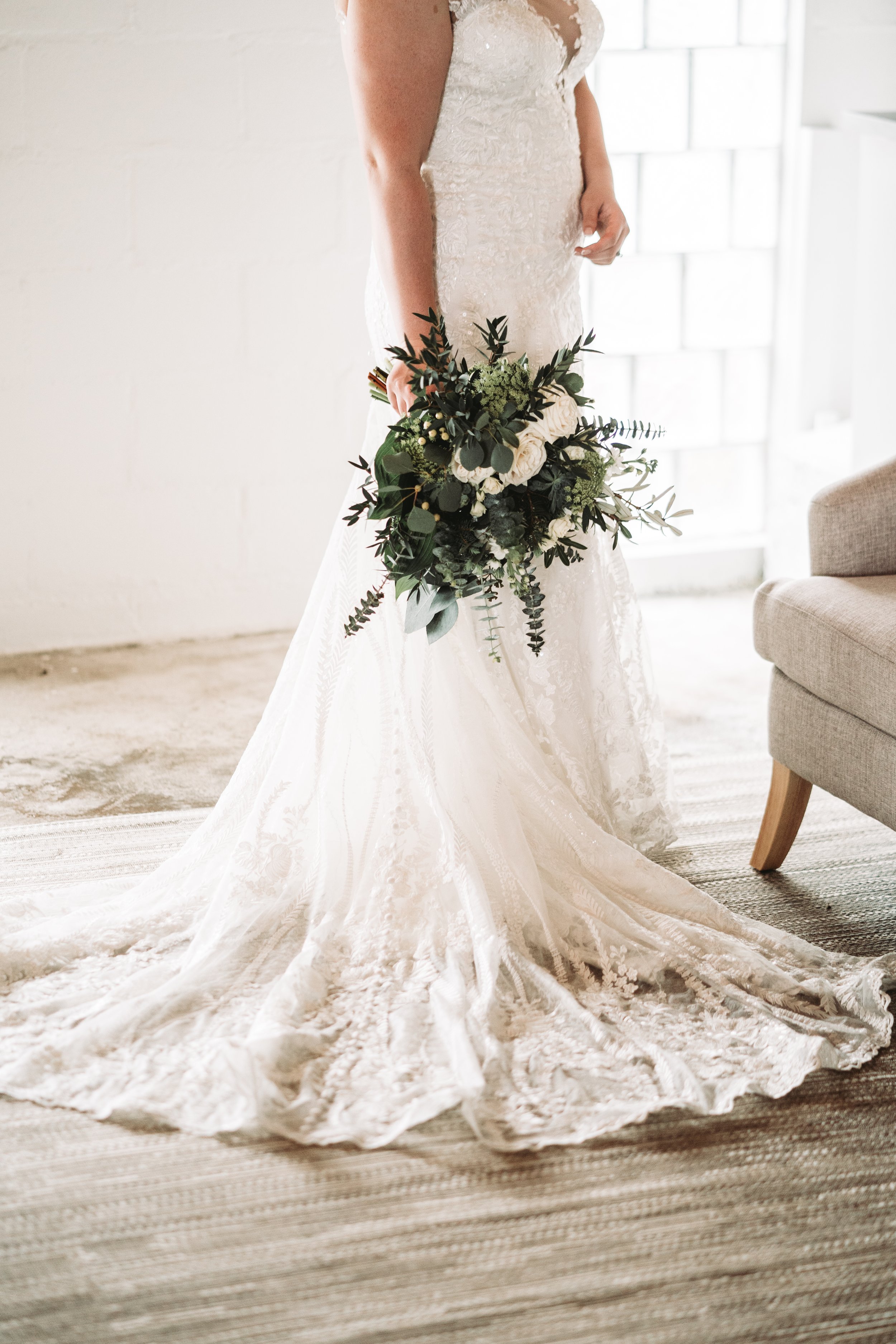 Bride in lace wedding dress holding a bouquet, standing indoors next to a chair.