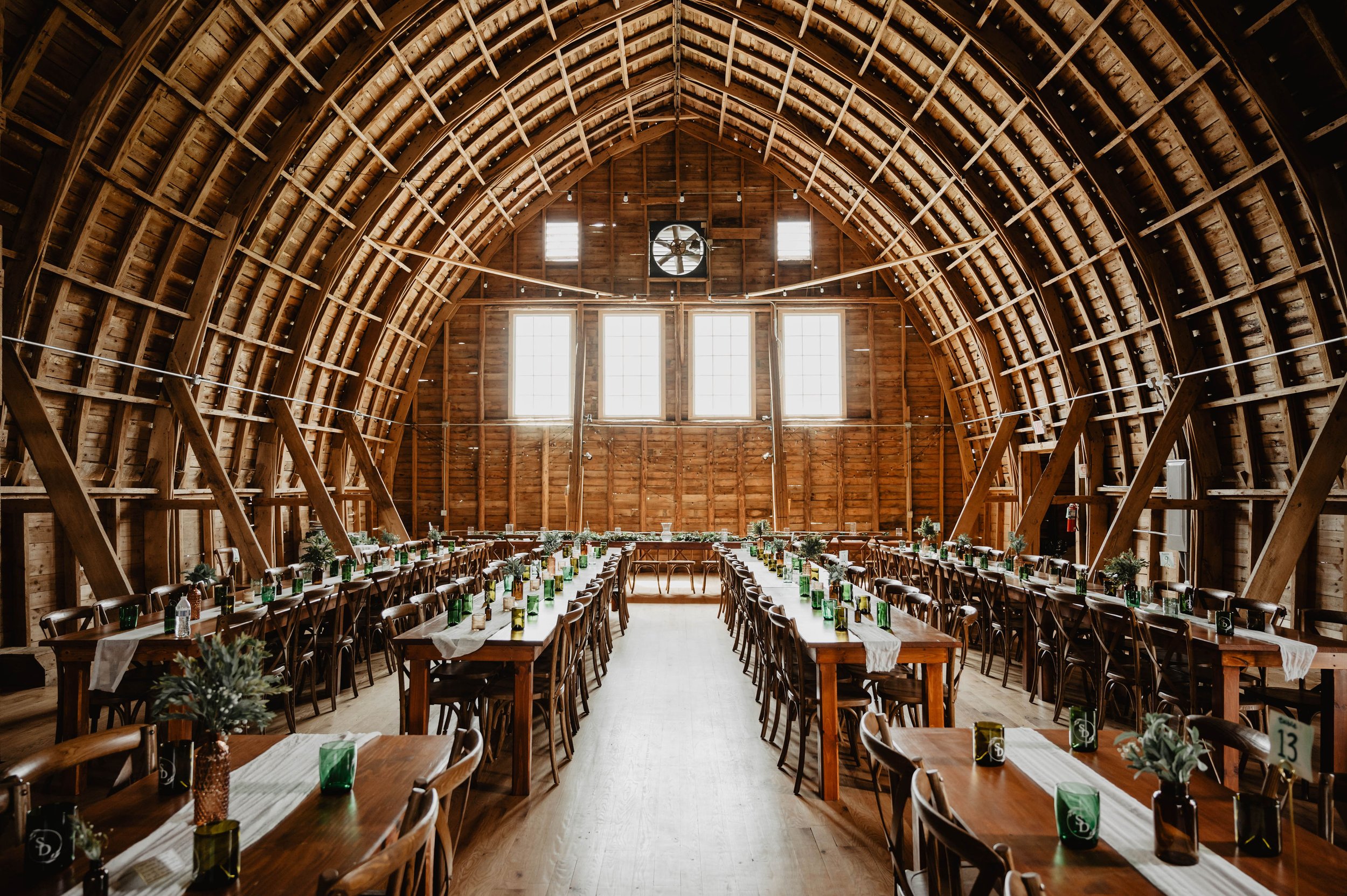 Interior of a rustic barn setup for an event, with long wooden tables, chairs, green glassware, and white table runners.