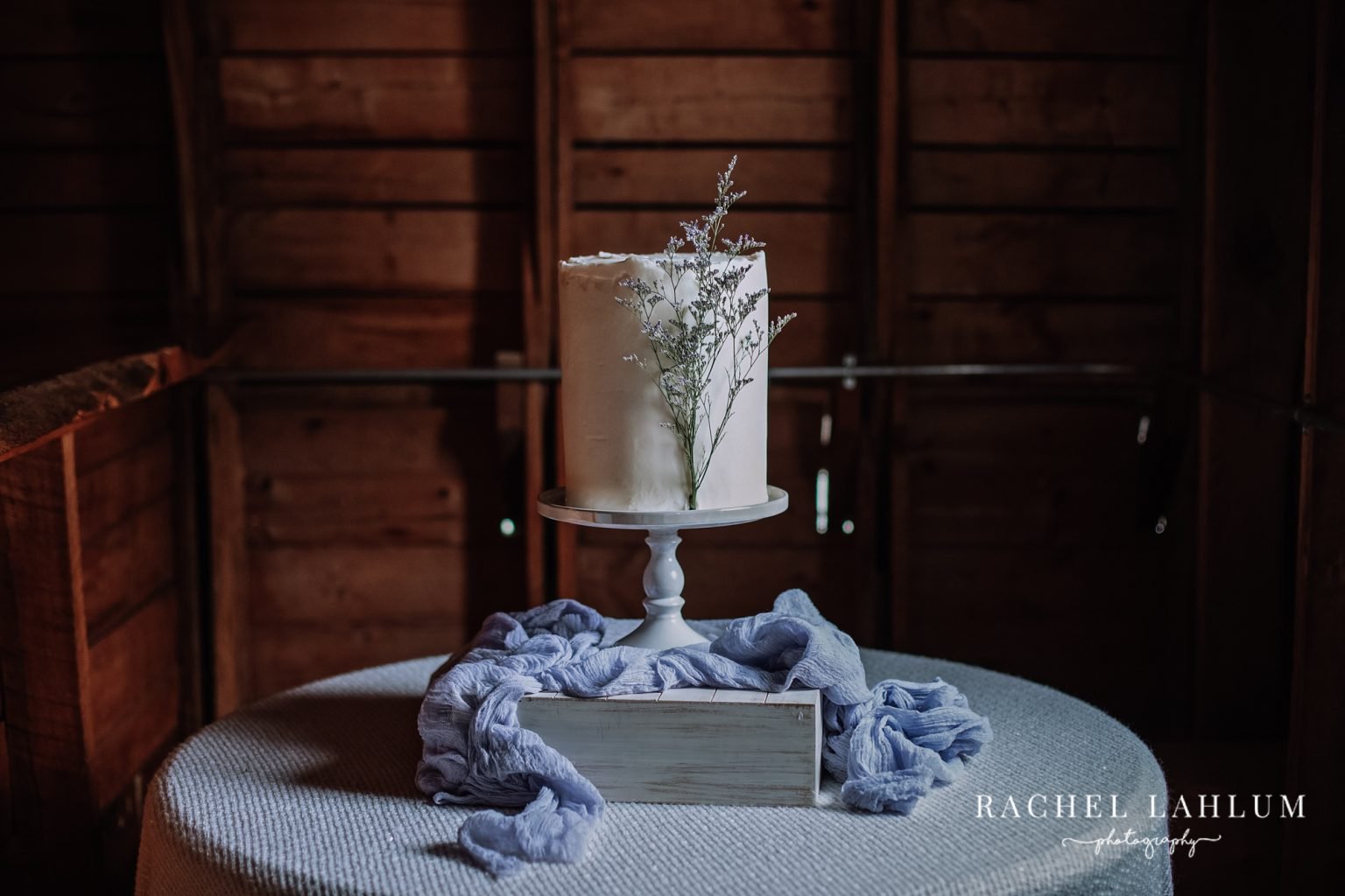 Minimalist white cake with floral decoration on a wooden box, draped with blue fabric, set on a round table in a rustic wood-paneled setting.