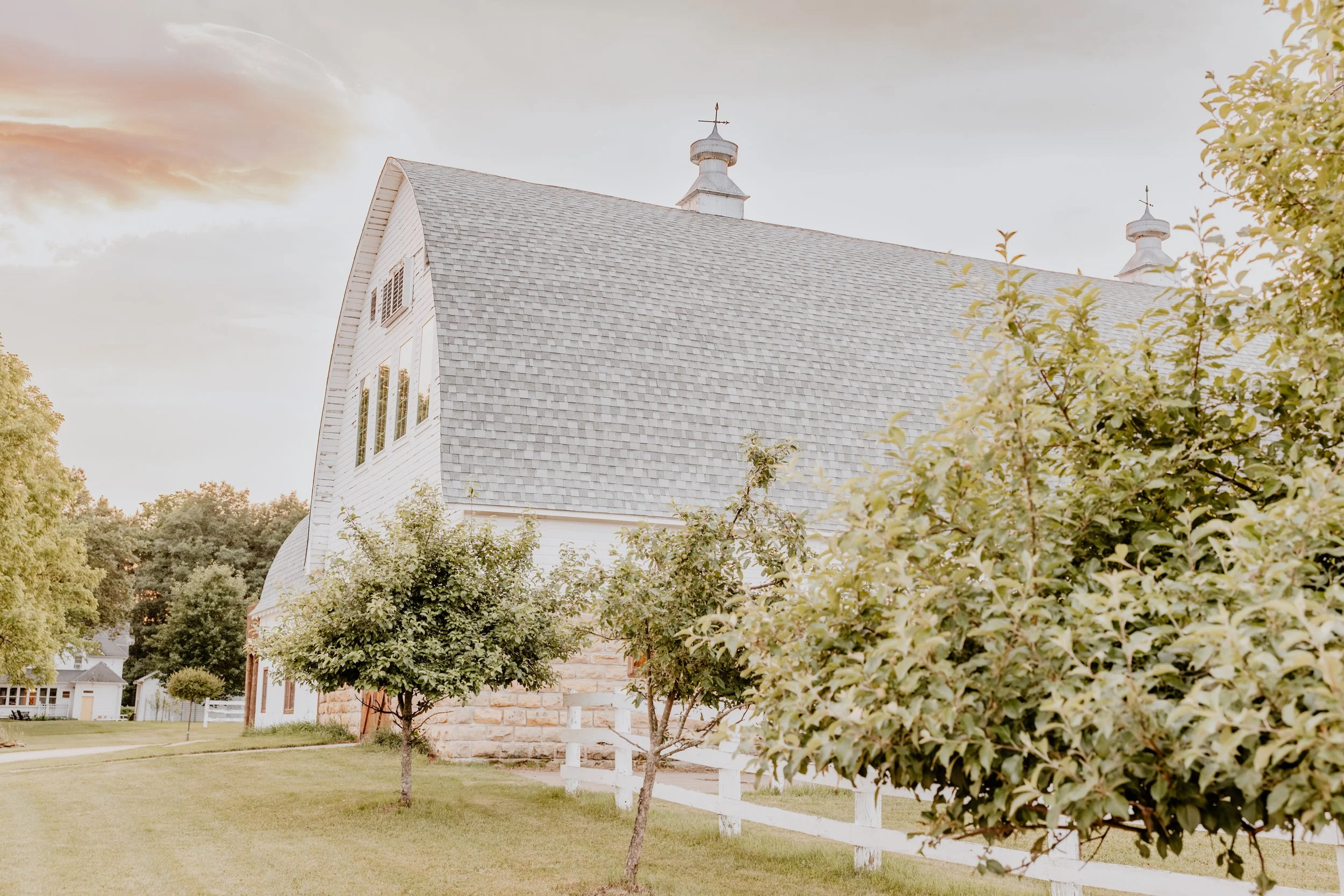 White barn with gray shingle roof, surrounded by green trees and grass, under a partly cloudy sky during sunset.