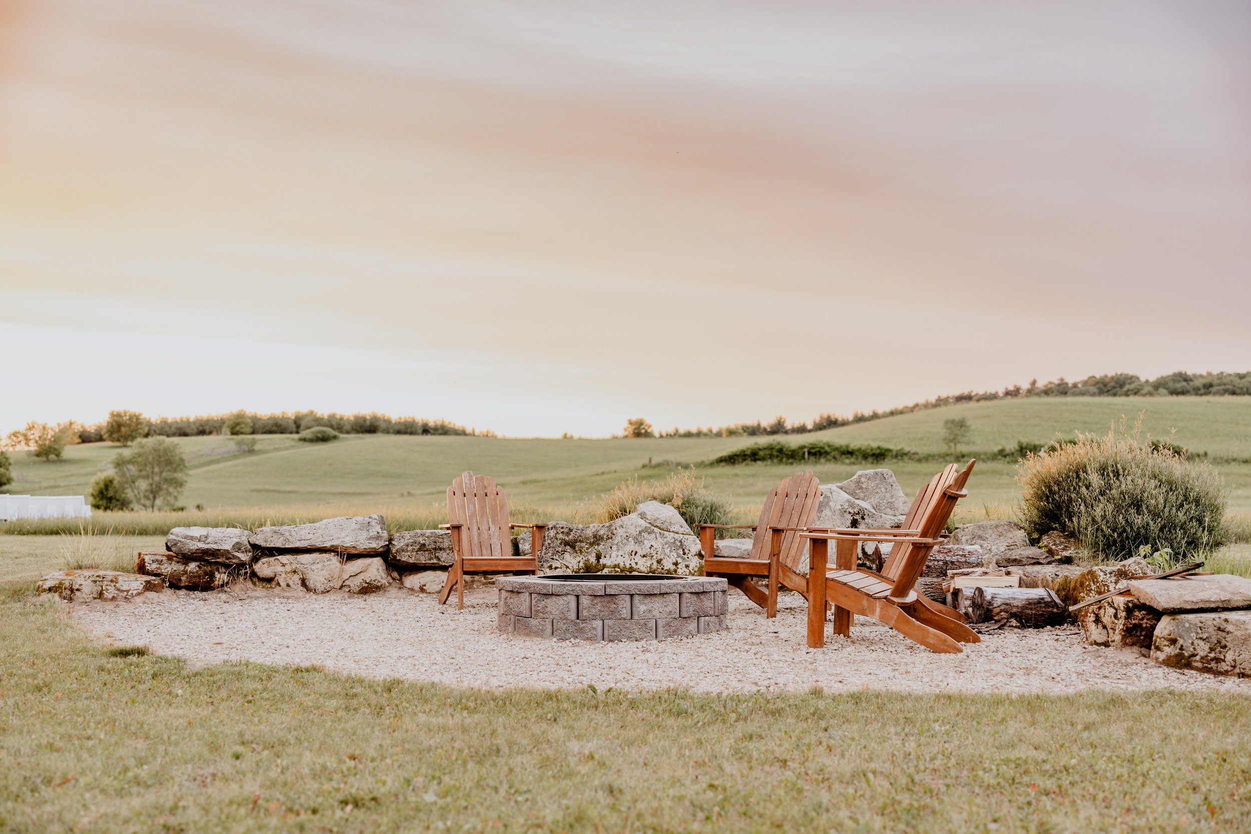 A cozy outdoor fire pit area with four wooden Adirondack chairs surrounding a stone fire pit, set in a spacious grassy landscape with gently rolling hills and trees in the background during sunrise or sunset.