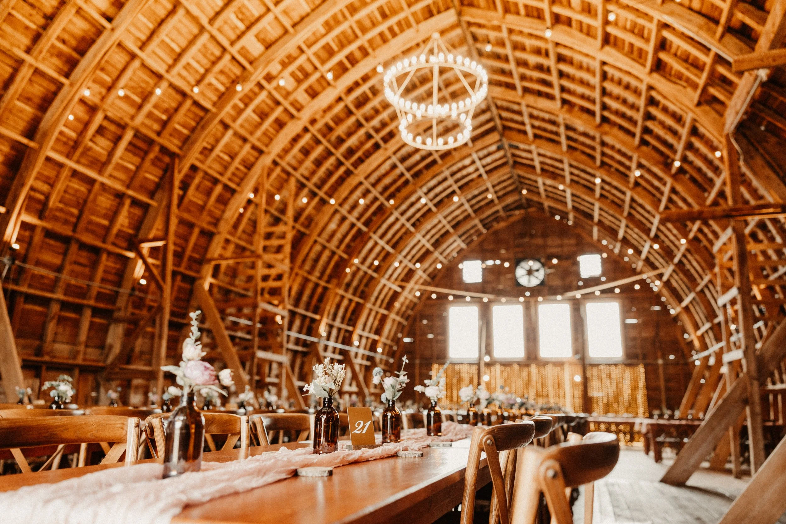 Long wooden banquet table inside a rustic barn with a high arched ceiling, decorated with small vases of flowers, string lights, and a central hanging chandelier.