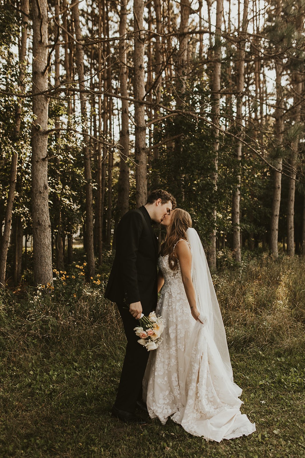 Bride and groom kissing in a forest setting, with the bride in a white lace gown and veil, and the groom in a black suit, surrounded by tall trees and wildflowers.