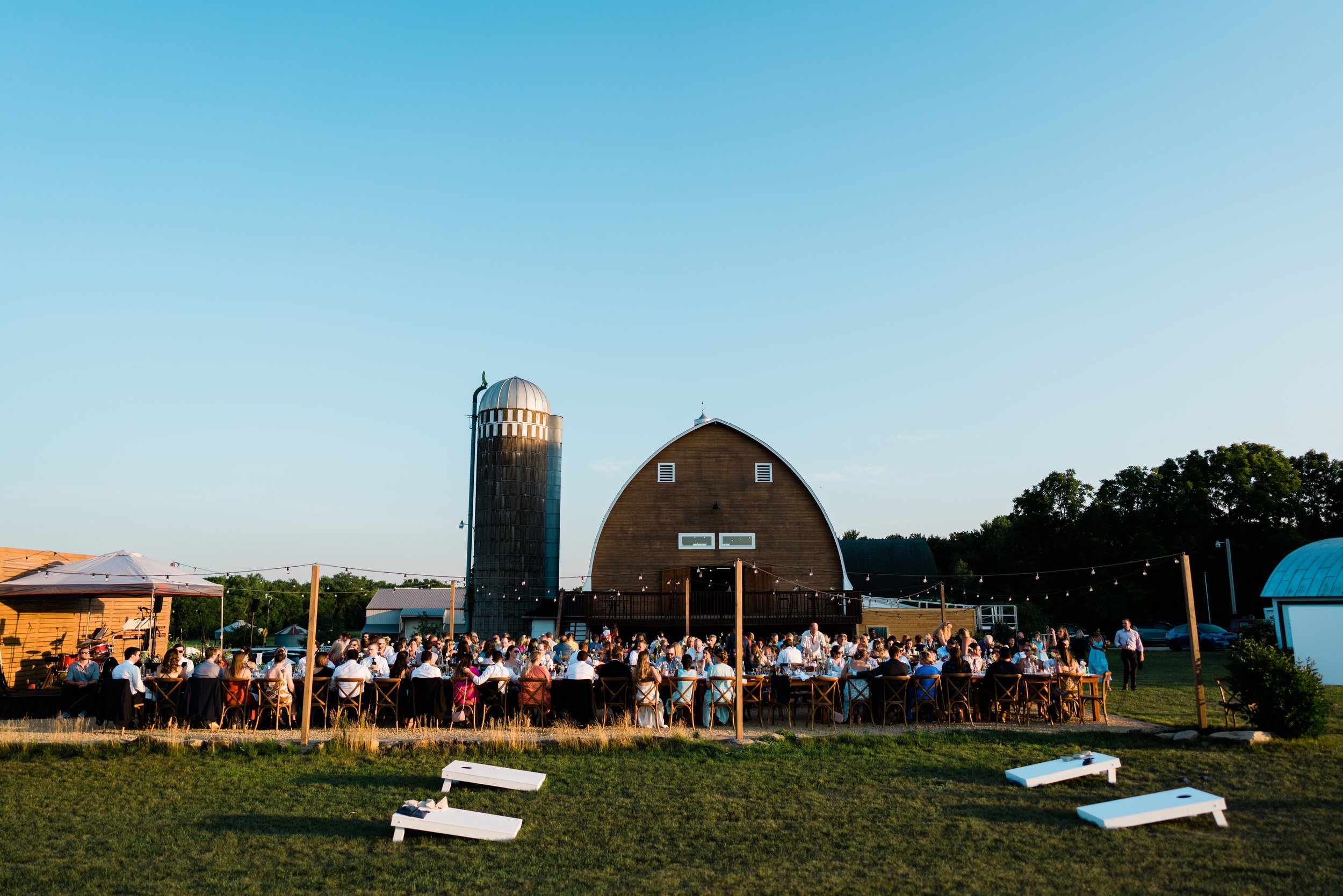 Outdoor gathering at a farm with people seated at long tables, barn and silo in background, blue sky, and cornhole game in foreground.