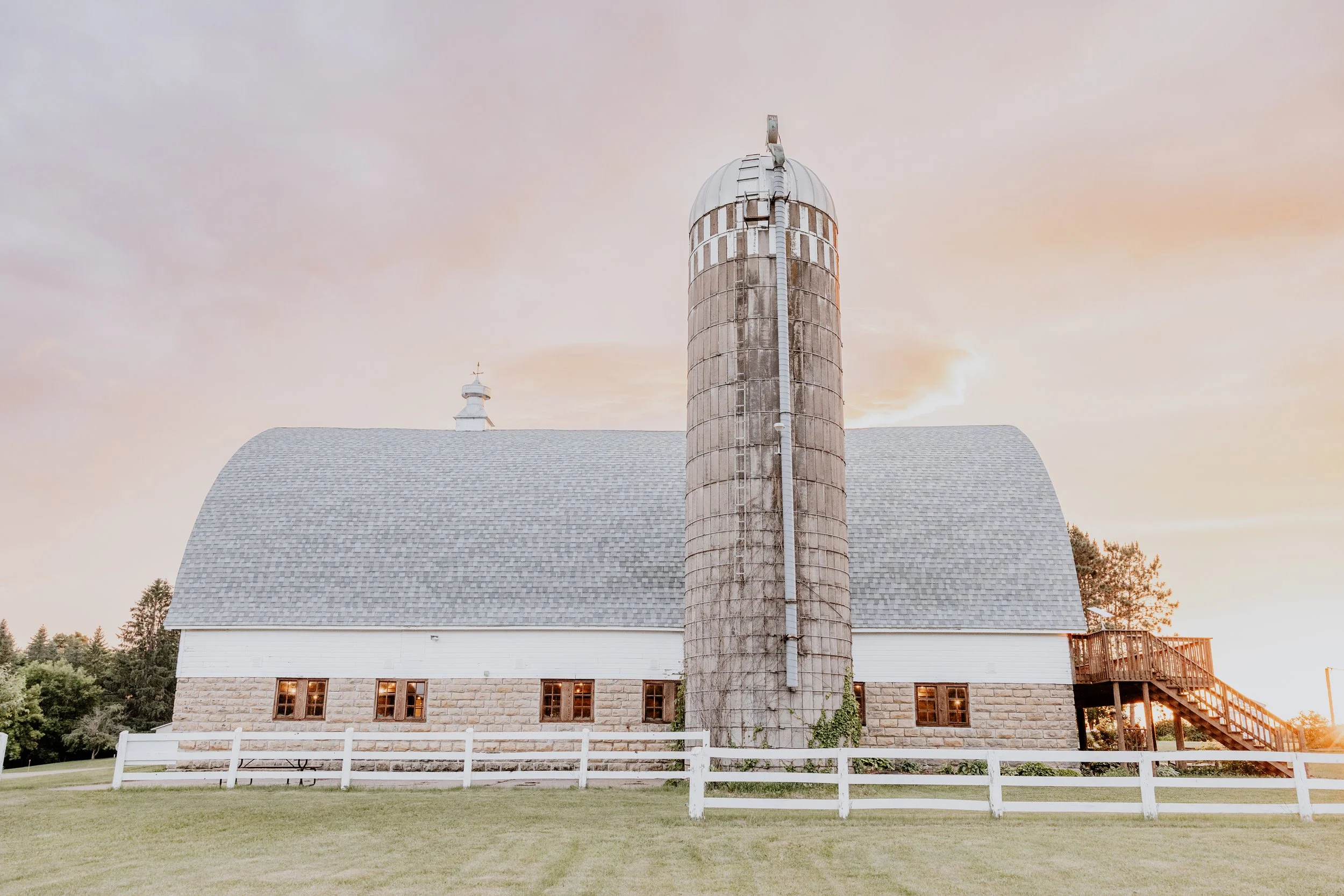 A white church with a stone foundation, dark roof, and a tall weathered silo in front, surrounded by a white fence and green grass, at sunset with a soft, pastel sky.