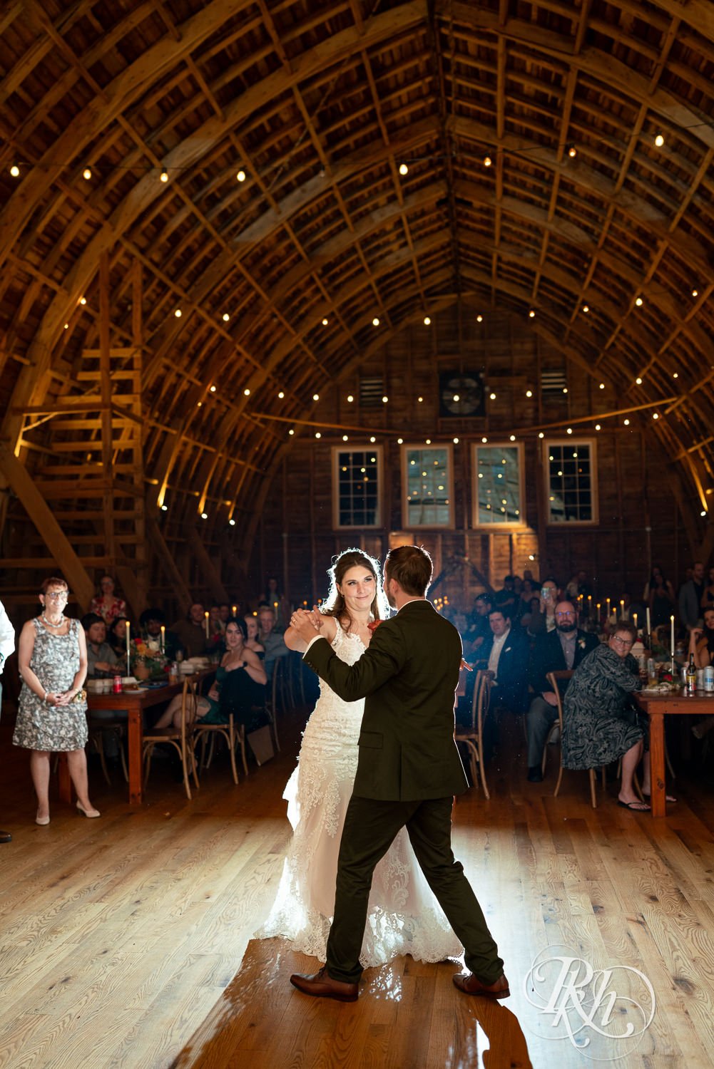 Bride and groom dancing in a rustic barn wedding venue with guests seated at tables, string lights hanging from the wooden ceiling.