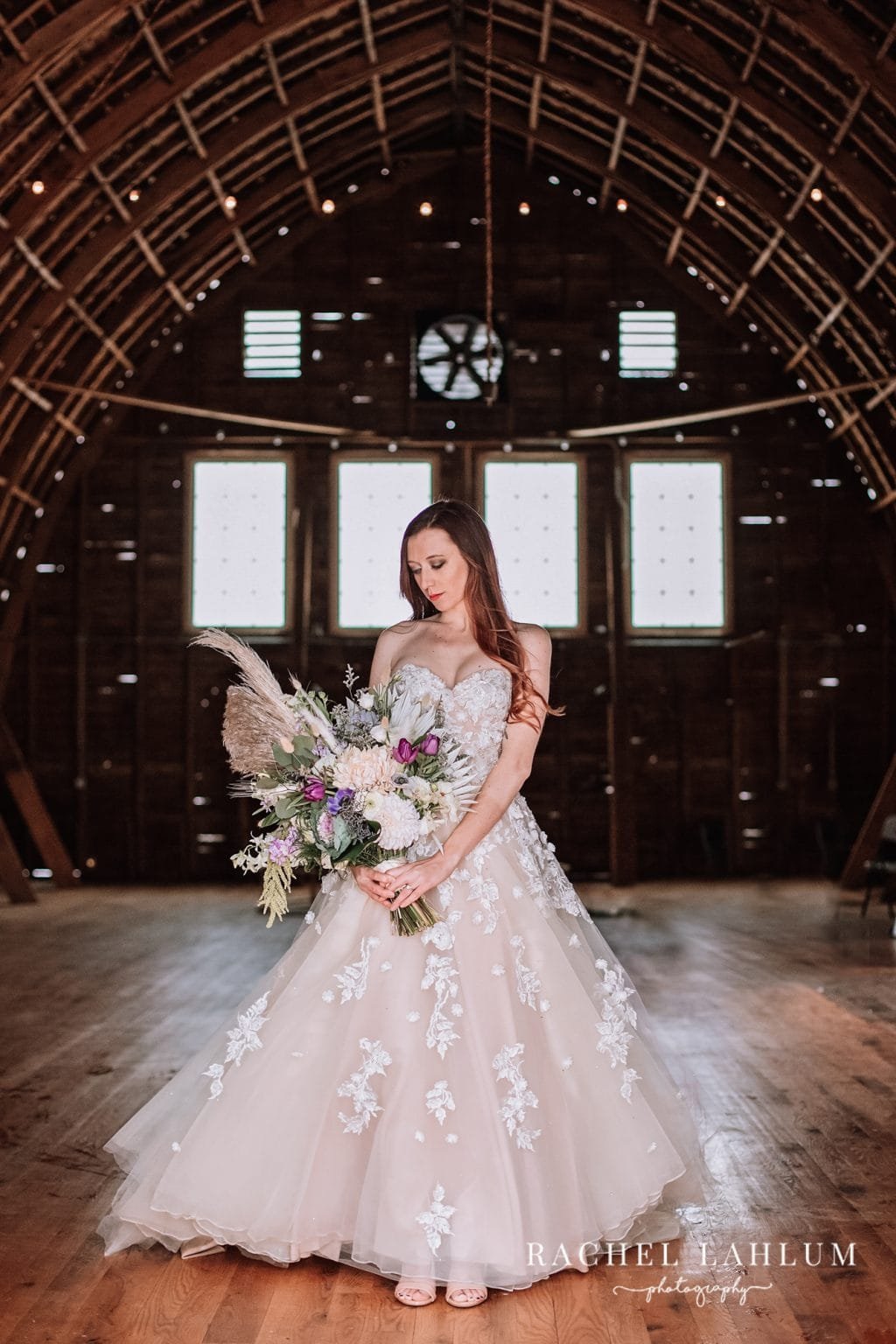 Bride in a floral wedding dress holding a bouquet inside a rustic barn.