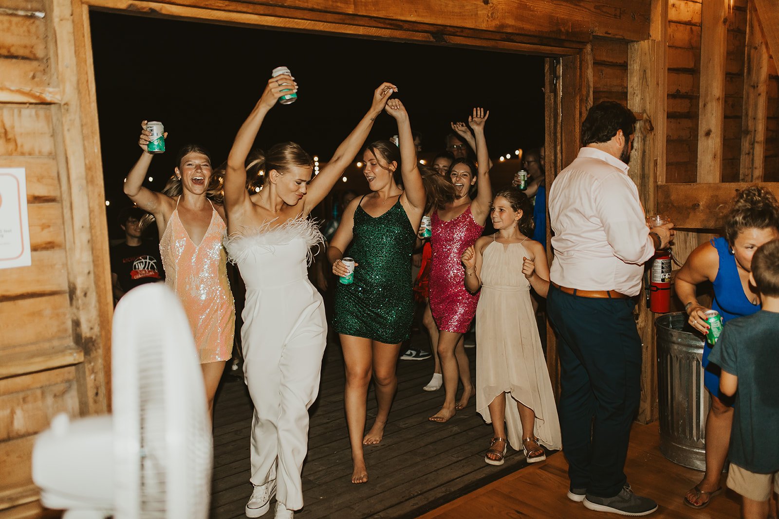 Group of people dancing and celebrating at a party, with women in colorful dresses holding drinks. The setting appears to be a wooden interior with warm lighting.