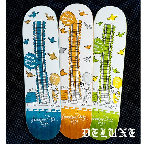 Three skateboard decks with firework-themed designs, each featuring the phrase "Support Your Local Skate Shop" and 'Skateboard Day 2024'. The decks are in blue, orange, and green, with illustrations of birds, buildings, and people.