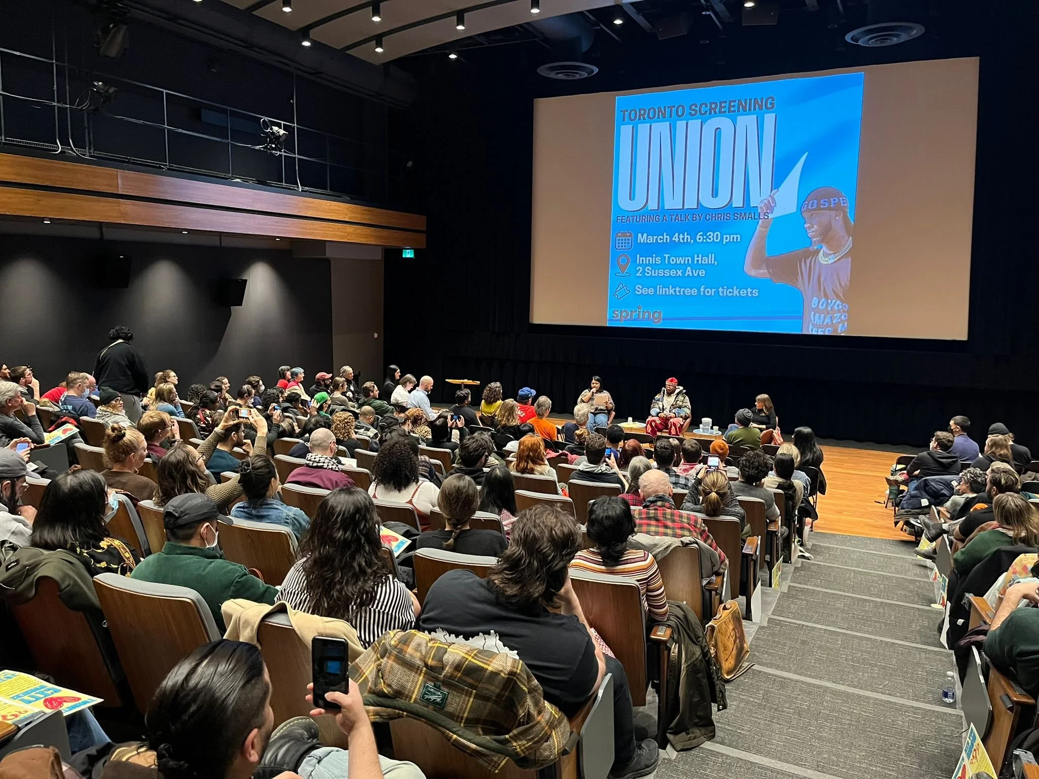 Audience seated in a theater attending a panel discussion on stage, with a large screen displaying an event titled 'Toronto Screening: Union' featuring Chris Smalls and director Brett Story, moderated by Sarah Shahid.