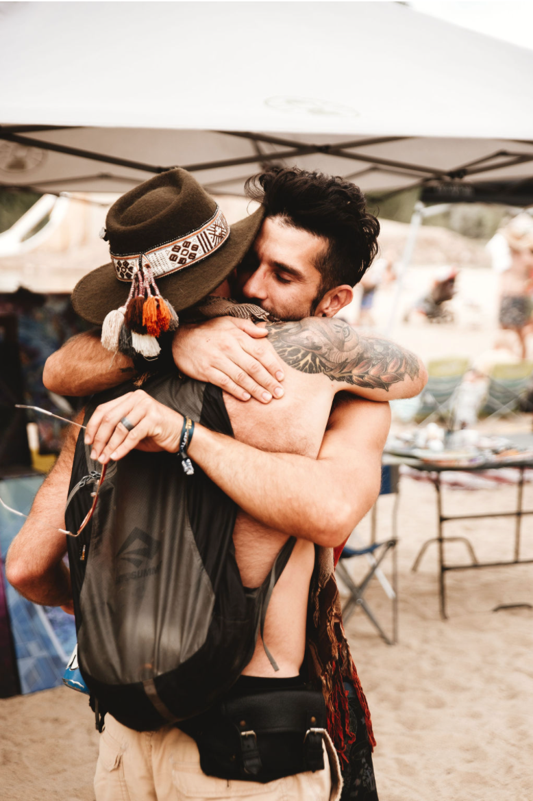 Two men embrace in a hug at an outdoor event, one wearing a wide-brimmed hat with colorful decorations, and the other with tattoos on his arm. The background features tents, chairs, and blurred people.