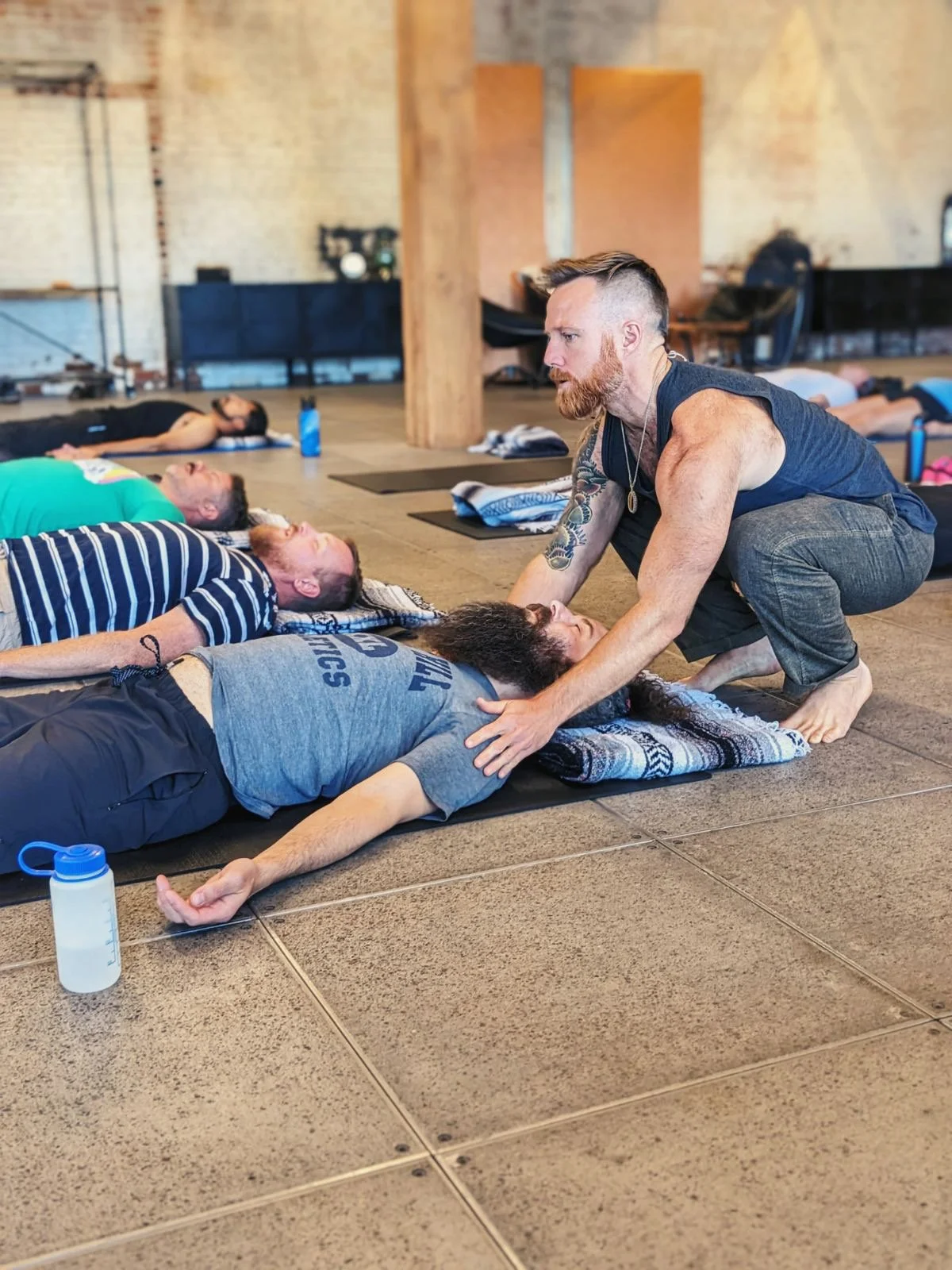 A group of people lying on yoga mats in a spacious studio, with an instructor assisting a person in a prone position with a stretching or healing practice.