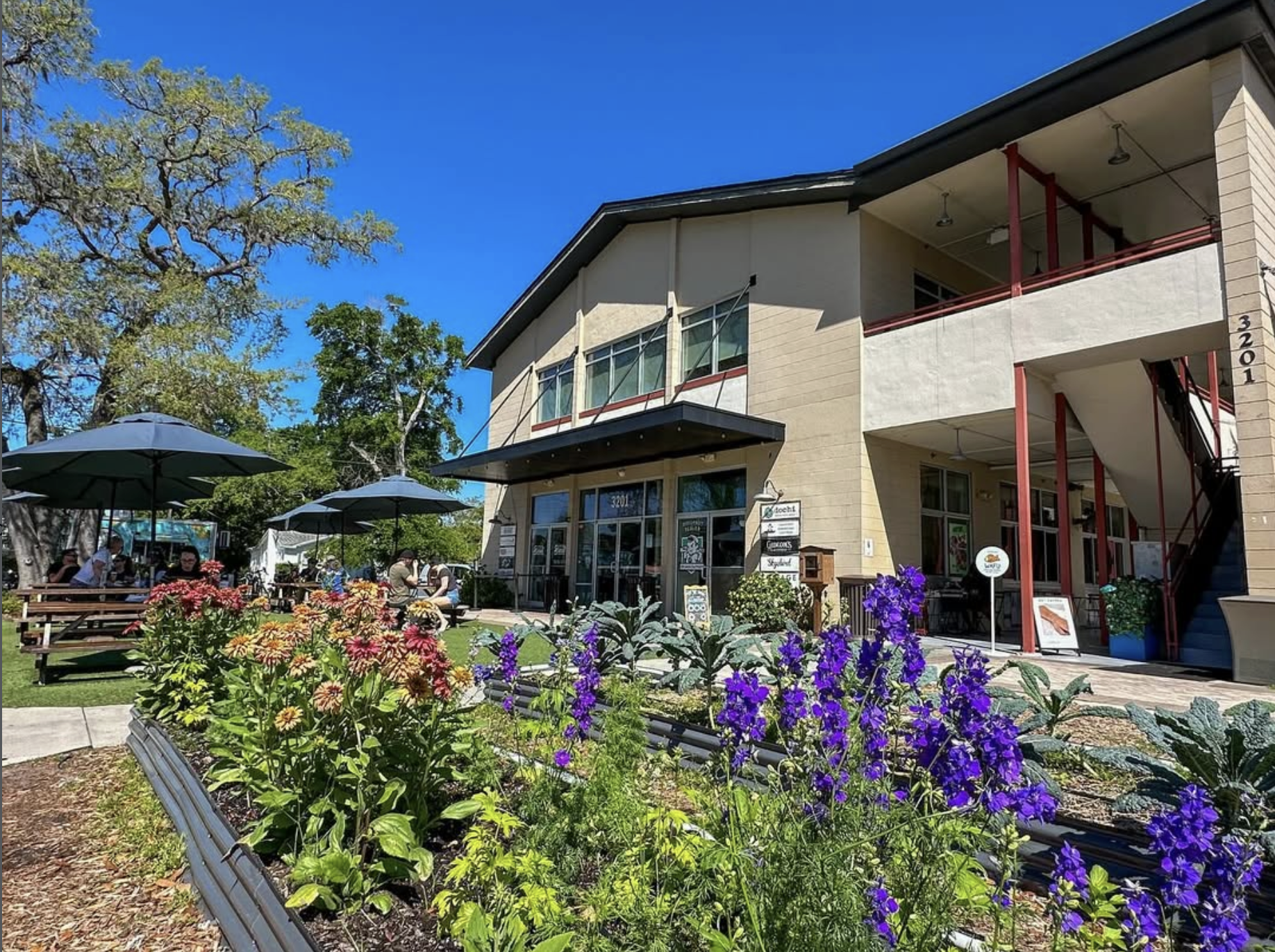 East End Market front entrance with outdoor picnic tables and raised planters with zinnias and delphiniums blooming in The APGD
