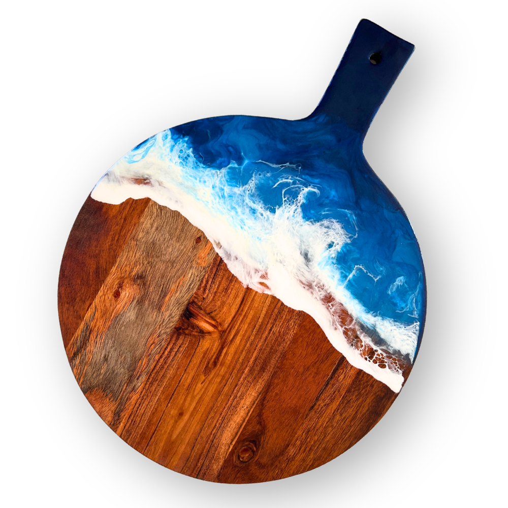 Round wooden serving board with ocean wave design painted on the top, featuring blue water, white foam, and a shoreline, with a black handle at the top.