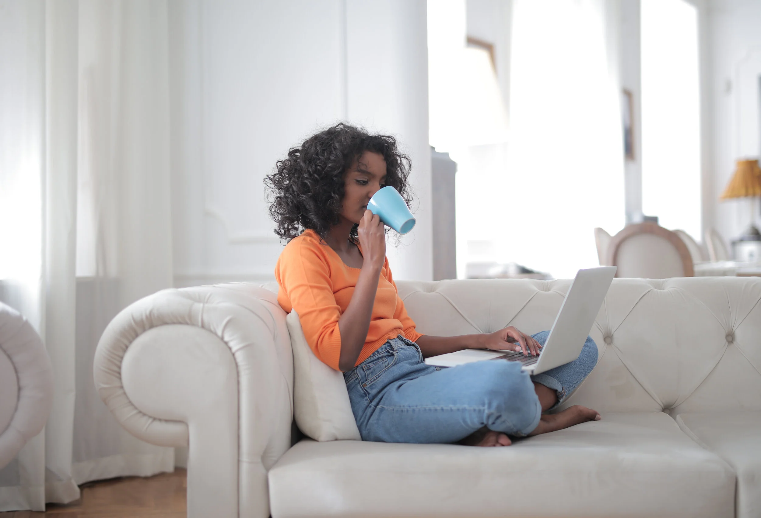 Canva - Positive young ethnic lady drinking hot beverage from cup while working on laptop and sitting on sofa at home.jpg