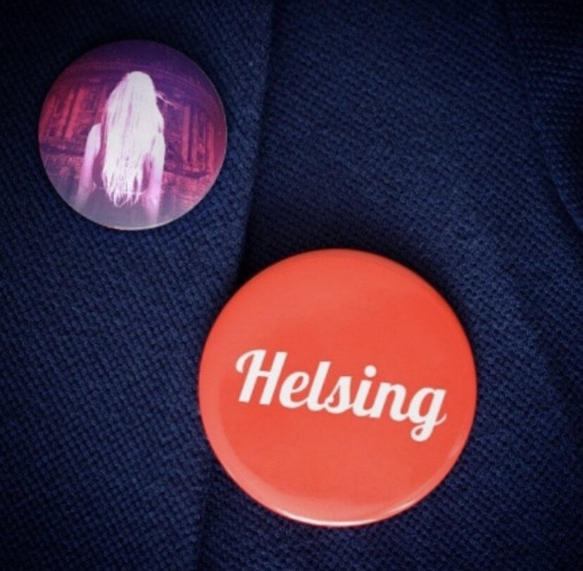 Two pins on a dark fabric; one features a woman with long hair in a pinkish-purple tone, the other reads "Helsing" in white text on a red background.