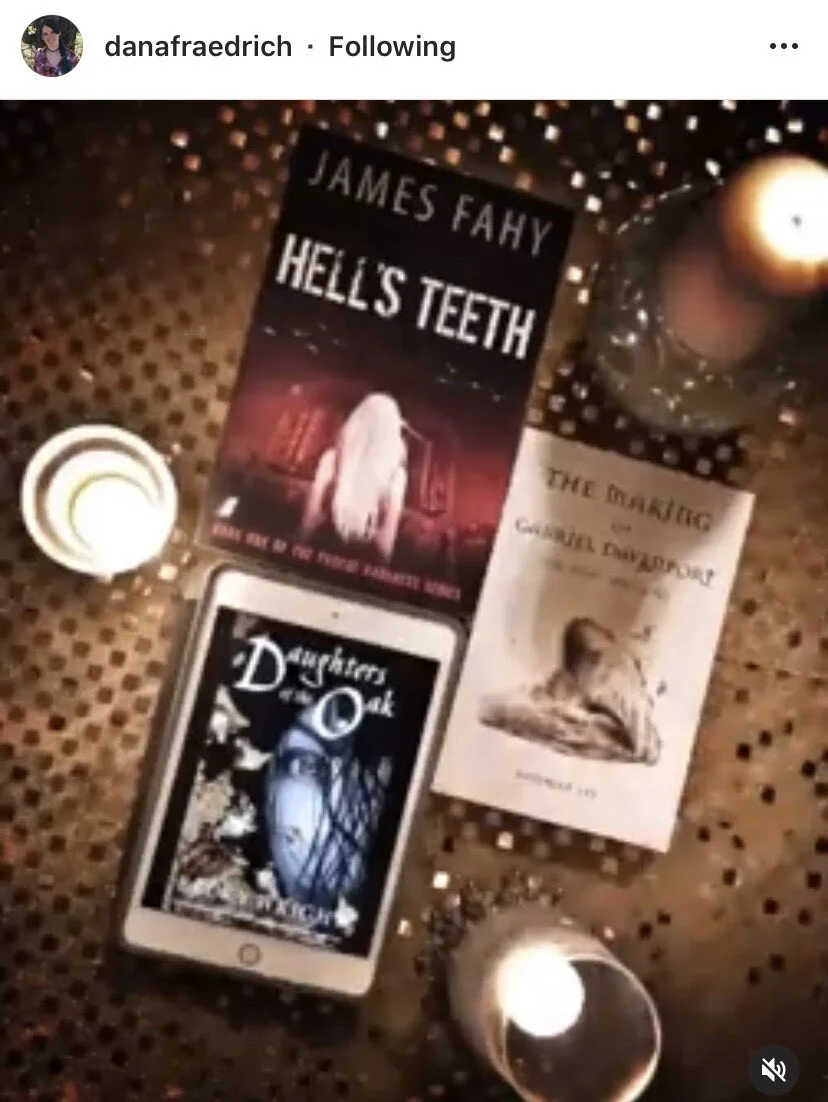 A book titled 'Hell's Teeth' by James Fahy, a deck of tarot cards titled 'Daughters of D'ark,' a book titled 'The Disabling' by Carole D. Williams, a smartphone showing the tarot card deck, and lit candles on a decorative surface.