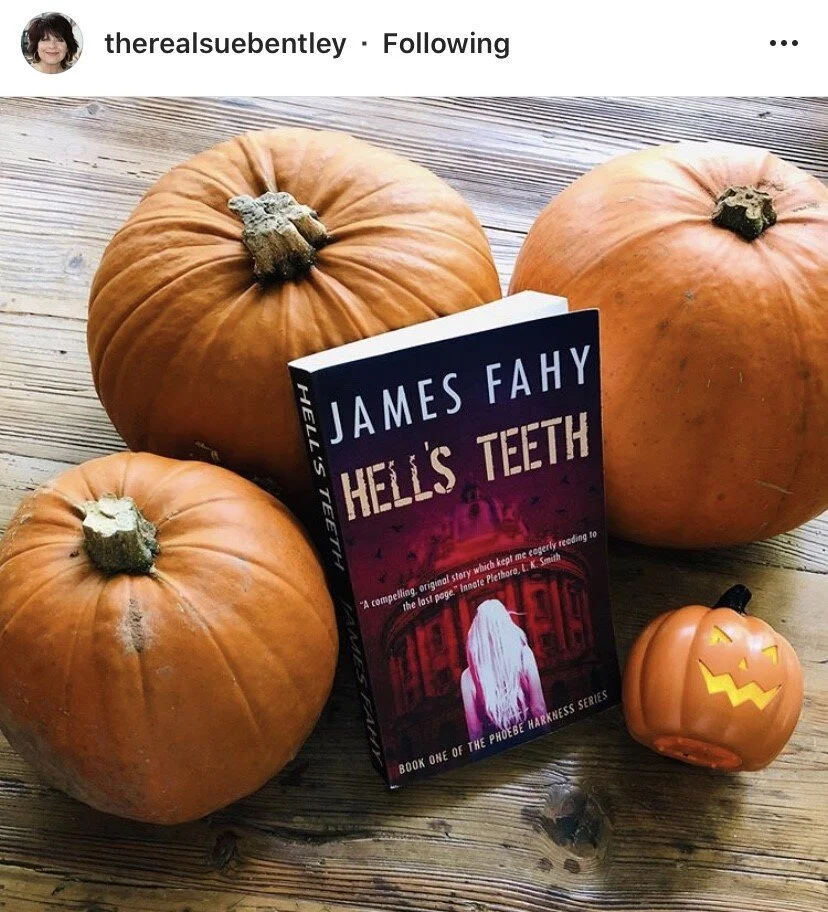 Three pumpkins, a Halloween-themed pumpkin container, and a book titled 'Hell's Teeth' by James Fahy on a wooden surface.