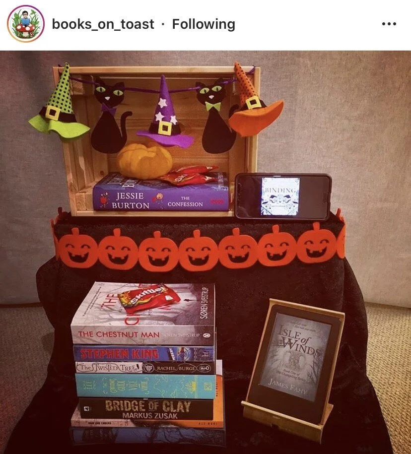 A Halloween-themed display with a wooden shelf, hanging felt witch hats, black cat cutouts, and a pumpkin on top. In front, there are several books, a stack of Halloween-themed books, a snack bag, a framed book cover, and a telephone showing part of 